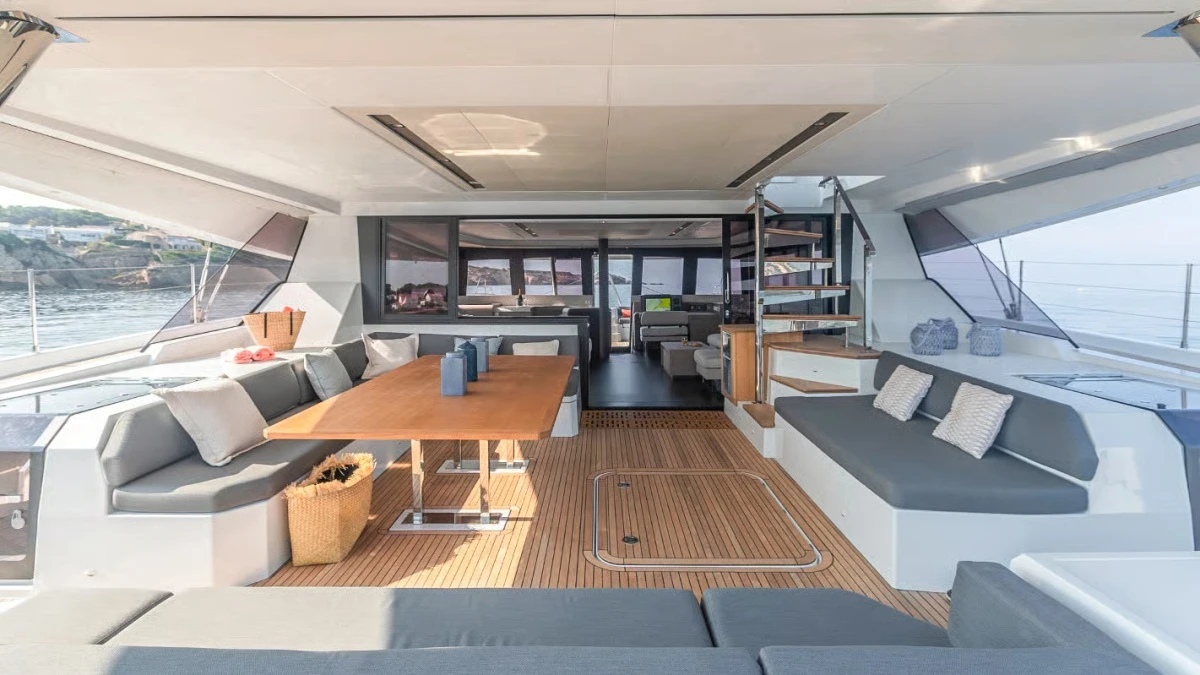 Fountaine Pajot Samana 59 katamaranın geniş arka güverte alanında, ahşap zemin ve gri minderli rahat oturma grupları, yemek masası ve cam kapılardan görünen şık iç salon ile mutfak bölümü, Singapur Yat Fuarı için tasarlanmış lüks bir konfor sunuyor.