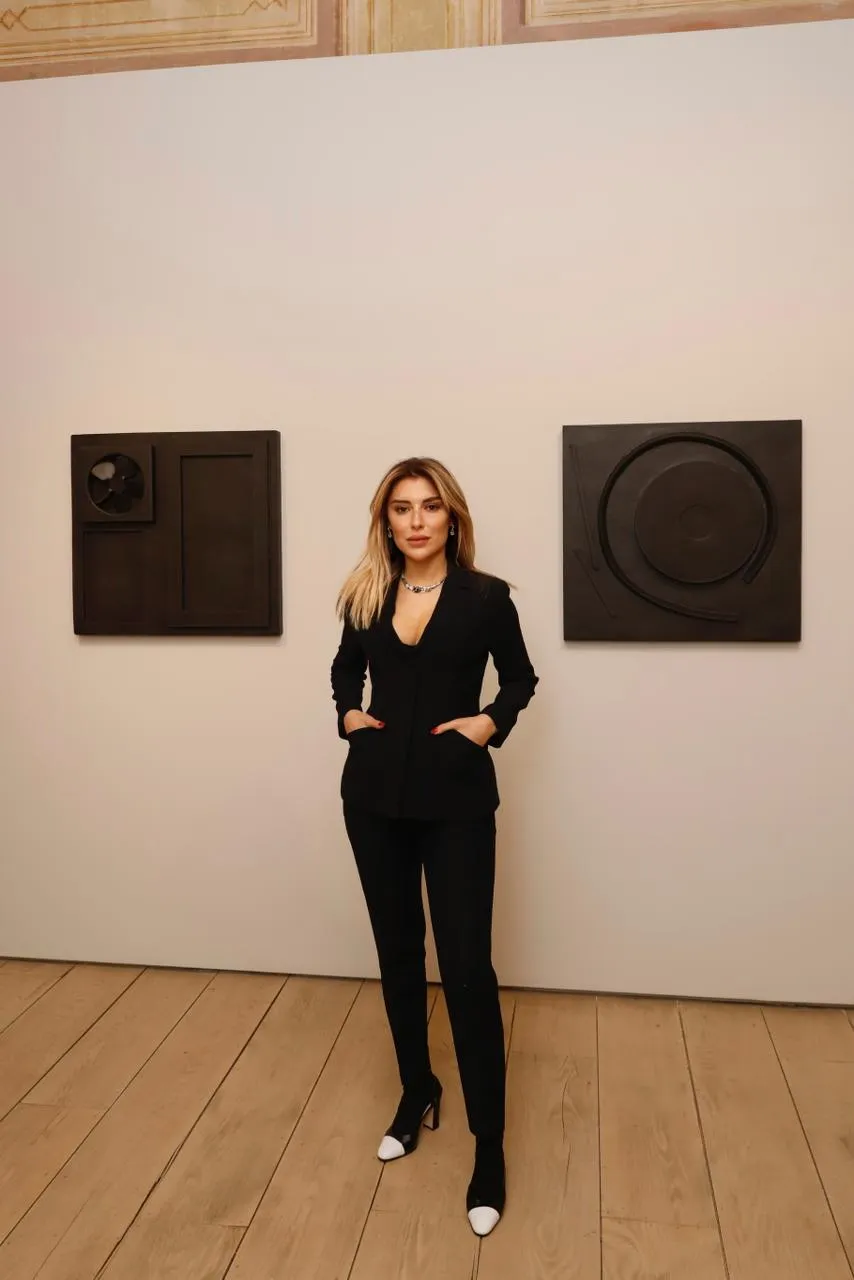 Sanat galerisindeki Avrupa II Sergisi'nde, sarı saçlı Sevil Dolmacı, siyah şık pantolon takımı, parlak kolyesi ve topuklu iki renkli botlarıyla, duvarda asılı koyu renkli iki modern soyut eserin önünde elleri cebinde poz veriyor.