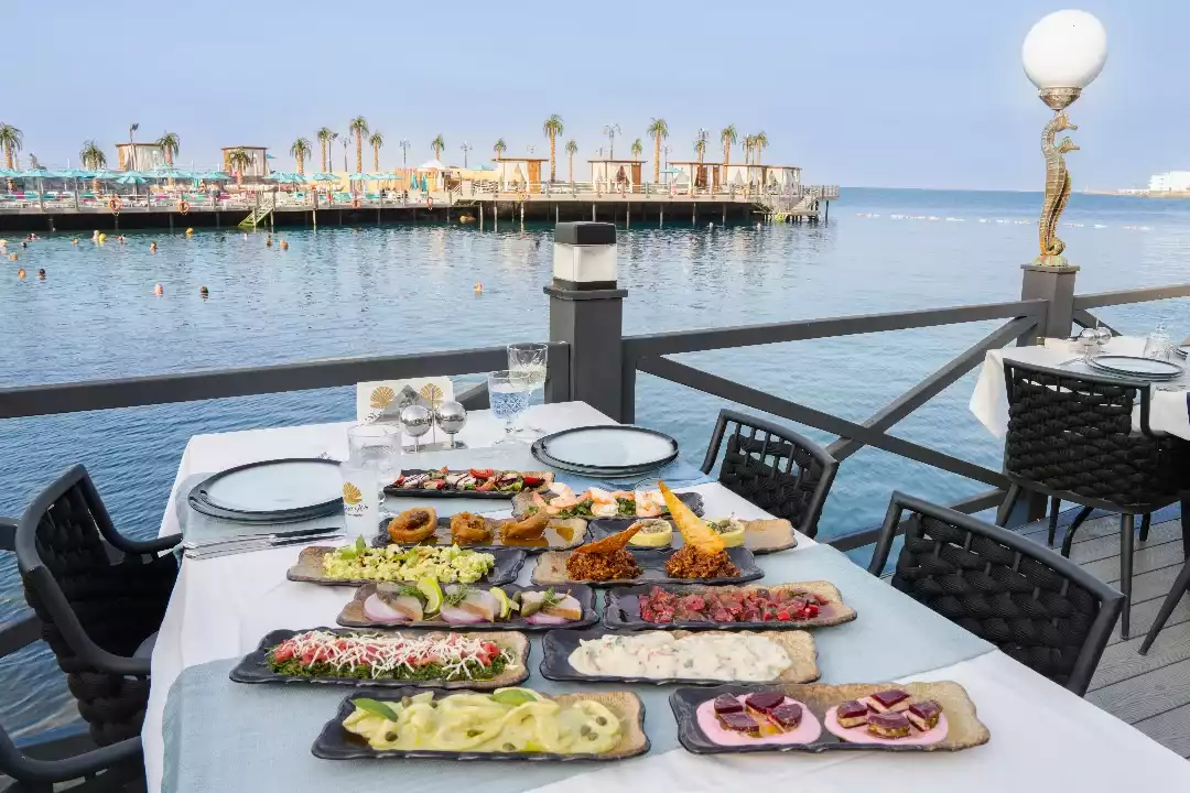 Deniz kenarındaki bir iskelede, lacivert denize nazır lüks bir restoranda beyaz örtülü uzun bir masanın üzerinde özenle hazırlanmış renkli ve çeşitli mezeler, salatalar ve küçük tatlılar sunuluyor; arka planda yüzen insanlar ve palmiye ağaçlarıyla dolu bir platform görünüyor.