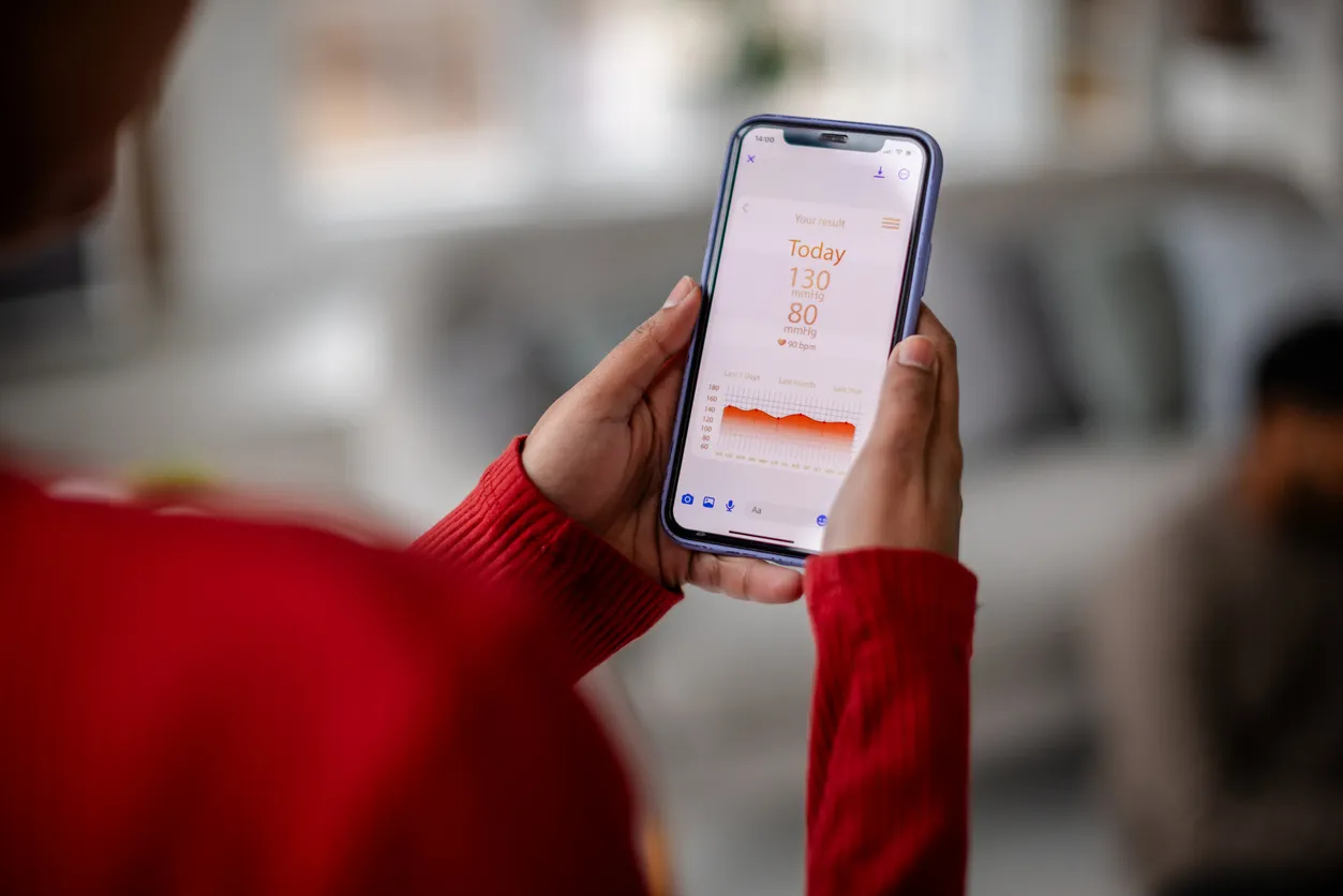 Kırmızı kazaklı bir kişi, elinde sağlık verilerini gösteren bir teknolojik alet olan akıllı telefonunu tutarken, ekranında 130/80 mmHg kan basıncı ve 90 bpm kalp atış hızı ile geçmiş 7 günlük ölçümleri gösteren turuncu bir grafik açıkça görülüyor, bu spor aktiviteleri sonrası takibi için ideal.