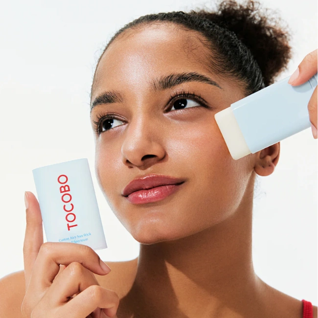 Beyaz arka planda, bir eliyle TOCOBO Cotton Airy Sun Stick ambalajını tutan ve diğer eliyle cildine aynı seriden bir güneş stick'i uygulayan genç bir kadın, ürünün pratik kullanımını ve pürüzsüz bitişini gösteriyor.