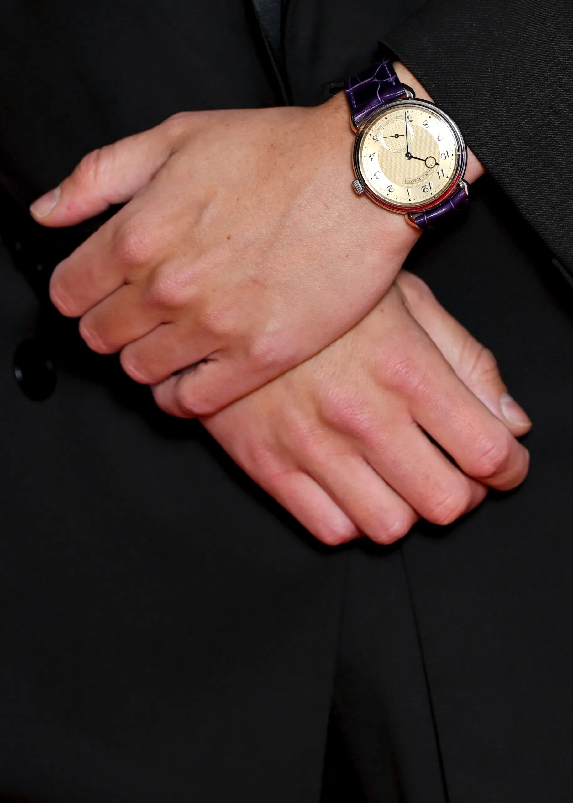 Timothée Chalamet'in şık ve dikkat çekici aksesuar seçimini vurgulayan bu yakın çekimde, koyu renk bir takım elbisenin kolları arasından çapraz duran elleri ve bileğindeki mor timsah derisi kayışlı, açık renk kadranında zarif Arap rakamları ve küçük saniye ibresi bulunan lüks bir kol saati görülüyor.