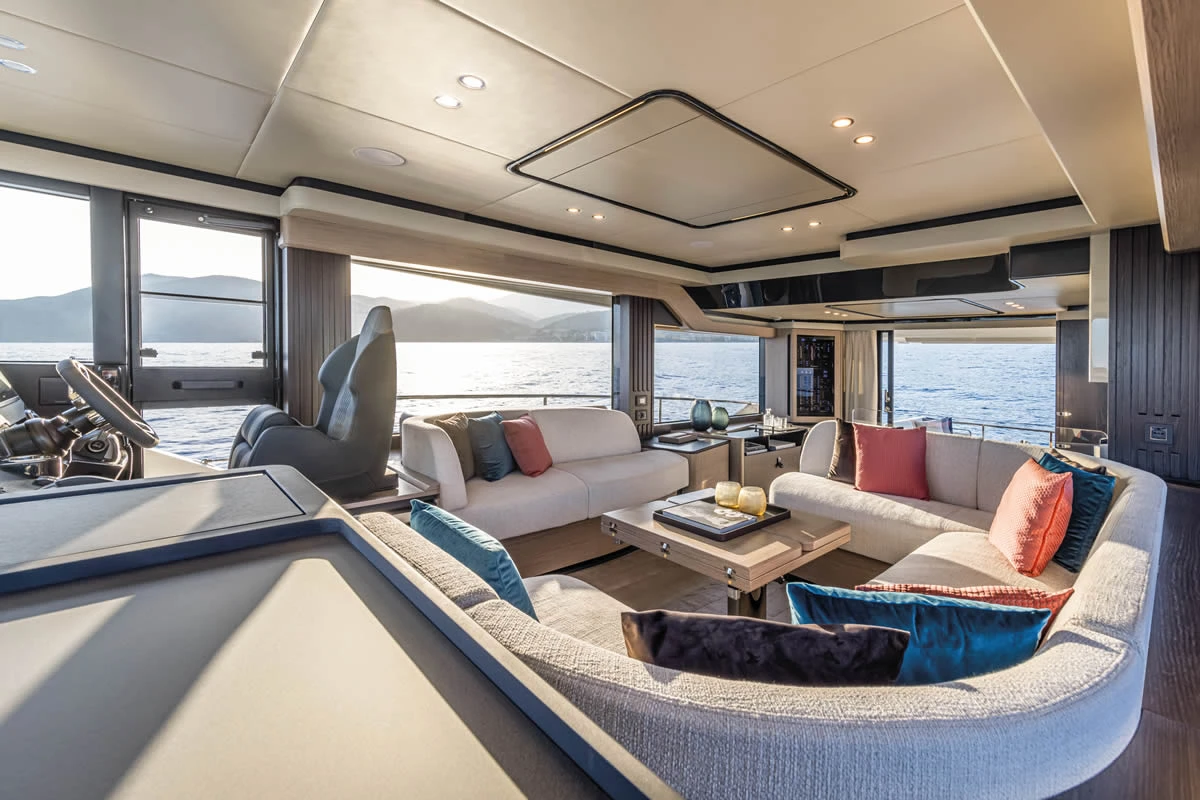Absolute Navetta 53'ün iç mekanında, geniş pencerelerden görünen parlayan deniz ve dağ manzarası eşliğinde, açık renkli konforlu kanepeler ve renkli minderlerle zenginleştirilmiş modern bir salon yaşam alanı dikkat çekiyor.