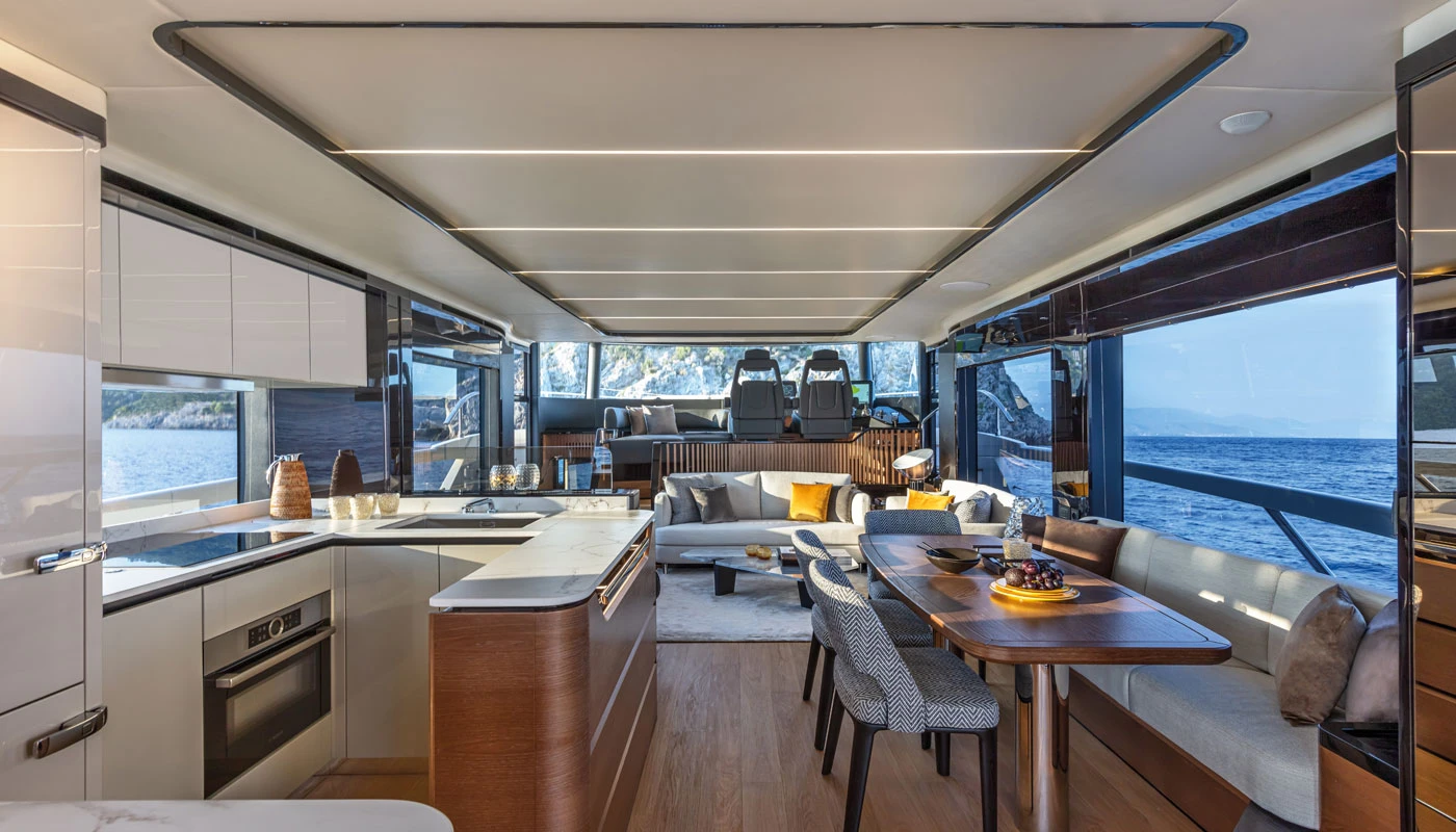 Absolute Navetta 64 yatının geniş ve lüks iç mekanında, modern tasarımlı beyaz mutfak bölümü, şık ahşap yemek masası ve konforlu salon alanı deniz manzaralı büyük pencerelerle birleşerek aydınlık ve ferah bir ortam sunuyor.