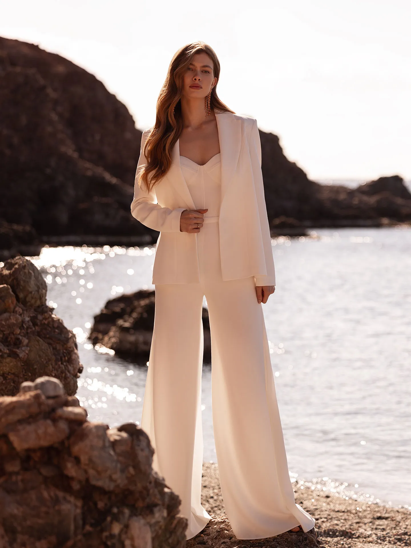 After party için elbise giymek istemeyen modern gelinlere ilham veren bu fotoğrafta, güneşin parladığı deniz kenarında beyaz bir blazer, straplez üst ve bol paça pantolondan oluşan şık bir takım elbise giyen genç bir kadın, uzun dalgalı saçları ve gösterişli küpeleriyle poz vererek düğün sonrası parti şıklığını alternatif bir tarzla vurguluyor.