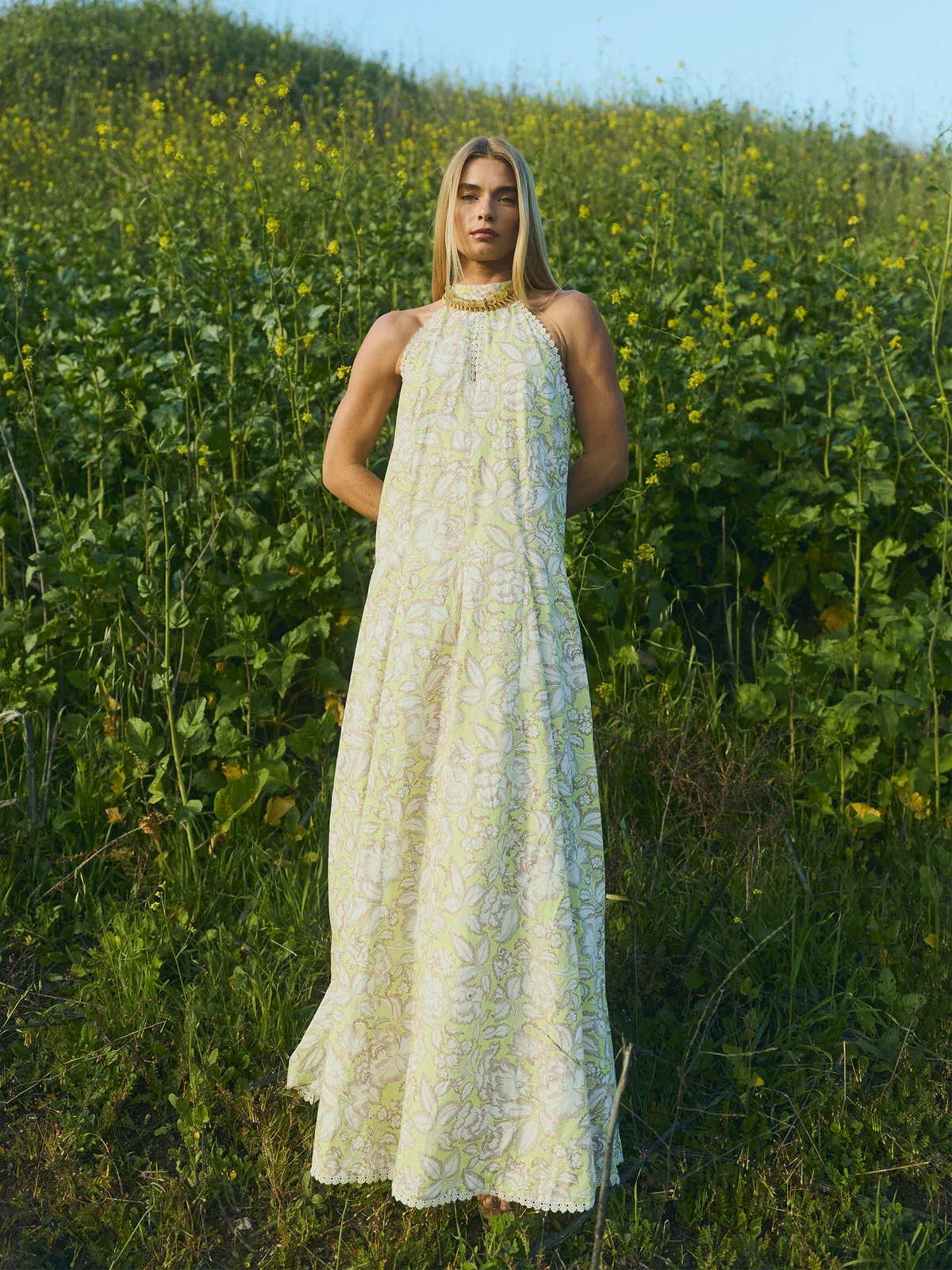 Kır düğünlerinin romantik atmosferine uygun, yemyeşil ve sarı çiçekli bir tarlada duran sarışın bir kadın, floral desenli, uçuk yeşil, halter yakalı, uçuş uçuş after party elbisesini gösterişli altın rengi bir kolye ile tamamlıyor.