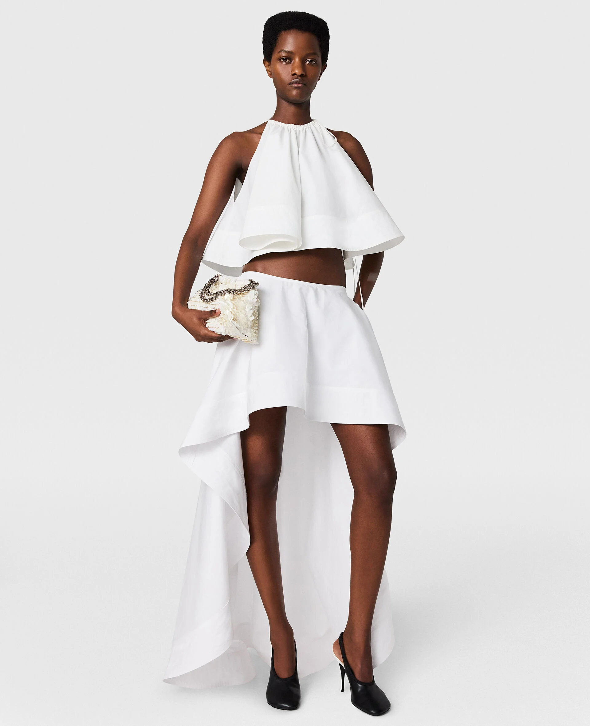 Model, Stella McCartney koleksiyonundan, enerjik ve rahat bir after party elbisesi olarak tercih edilebilecek, boyundan bağlamalı, katmanlı beyaz crop top ile önü kısa, arkası uzun, heykelsi silüete sahip asimetrik eteği bir arada giyerek dans pistinde özgürce hareket edebilen modern gelinlere ilham veriyor.