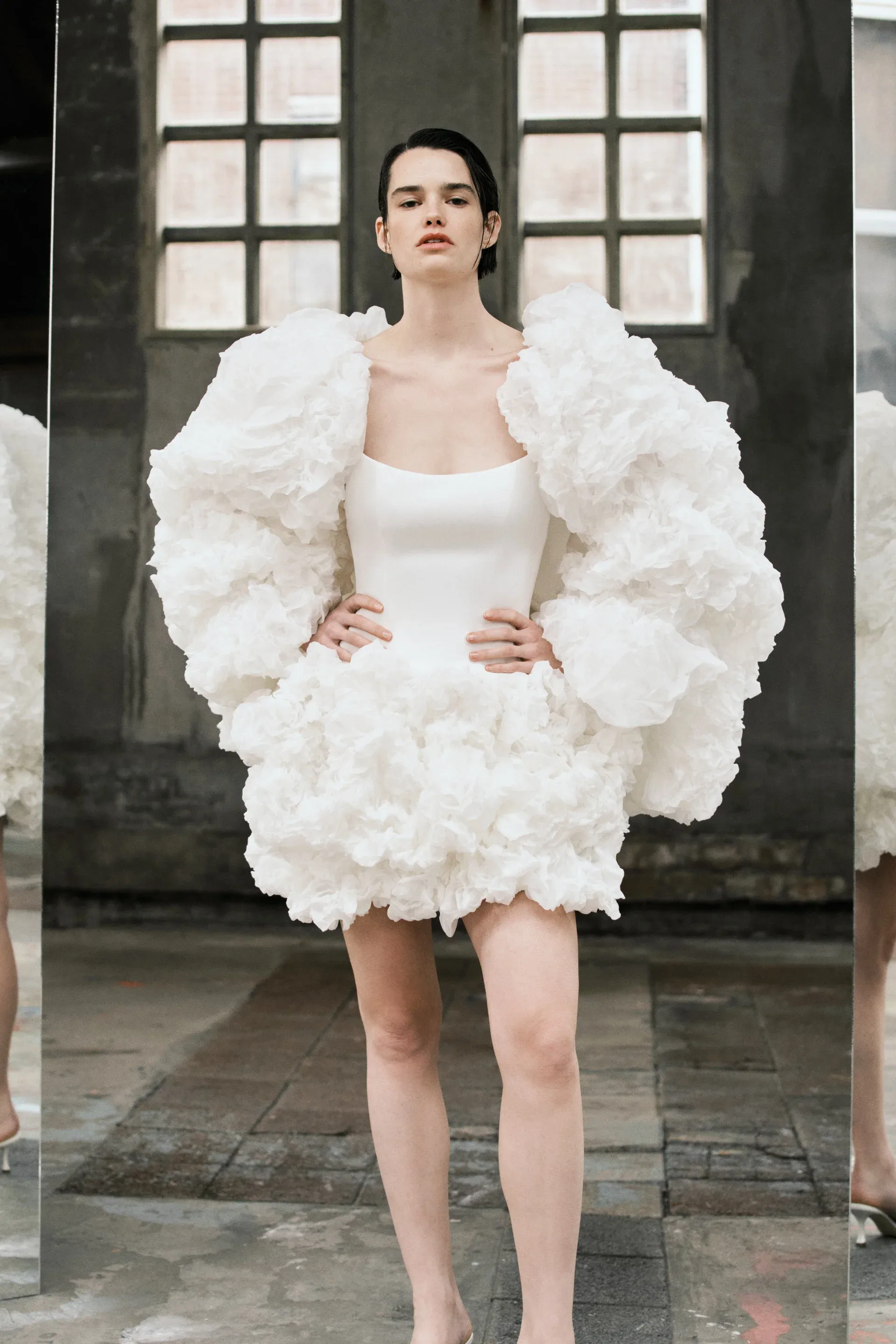 Bir model, şık ve modern bir after party elbisesi giymiş, Victor & Rolf tasarımı beyaz, hacimli fırfırlı kollara ve etek kısmında üç boyutlu çiçek formlarına sahip mini elbise, gecenin enerjisine eşlik etmek isteyen gelinler için göz alıcı bir seçenek sunuyor.