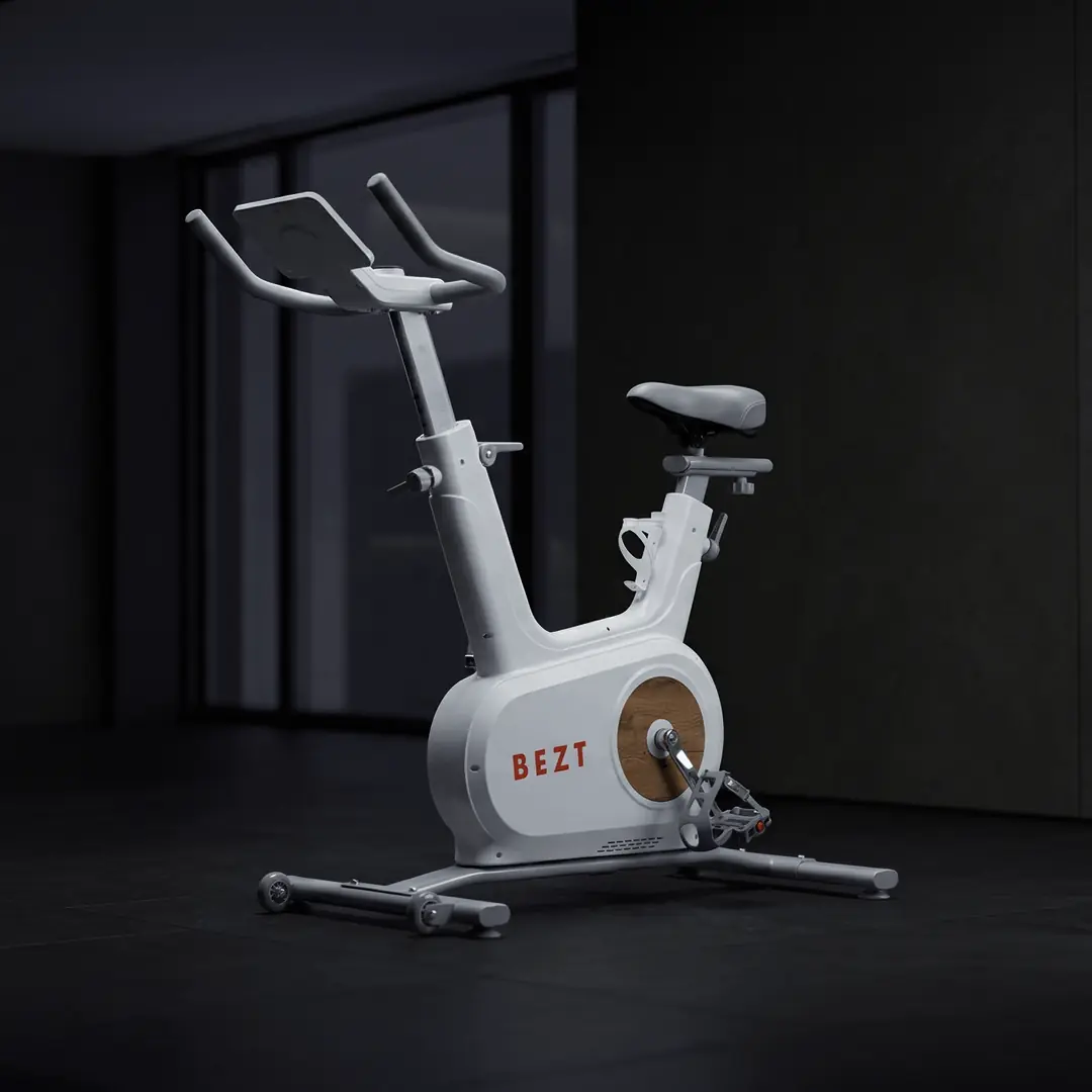 Şık ve modern tasarımlı, beyaz renkteki BEZT marka akıllı fitness bisikleti, gri selesi, ahşap görünümlü tekerlek kaplaması ve dijital ekran paneli için ayrılmış geniş gidonlarıyla koyu zeminli bir iç mekanda, geniş pencerelerin hafifçe yansıdığı bir ortamda sergileniyor.