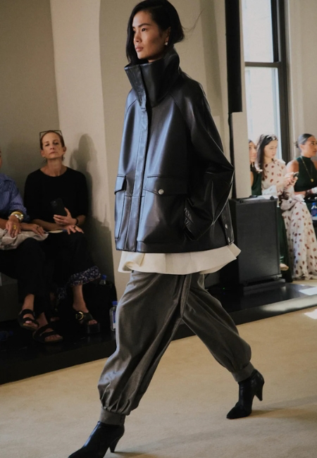 Altuzarra 2026 İlkbahar-Yaz podyumunda, koyu renkli, yüksek yakalı, oversize deri görünümlü bir ceket ve paçaları lastikli, hacimli gri balon pantolon giyen model, yeni ve güçlü bir siluetle adeta bir moda akımına öncülük ediyor.