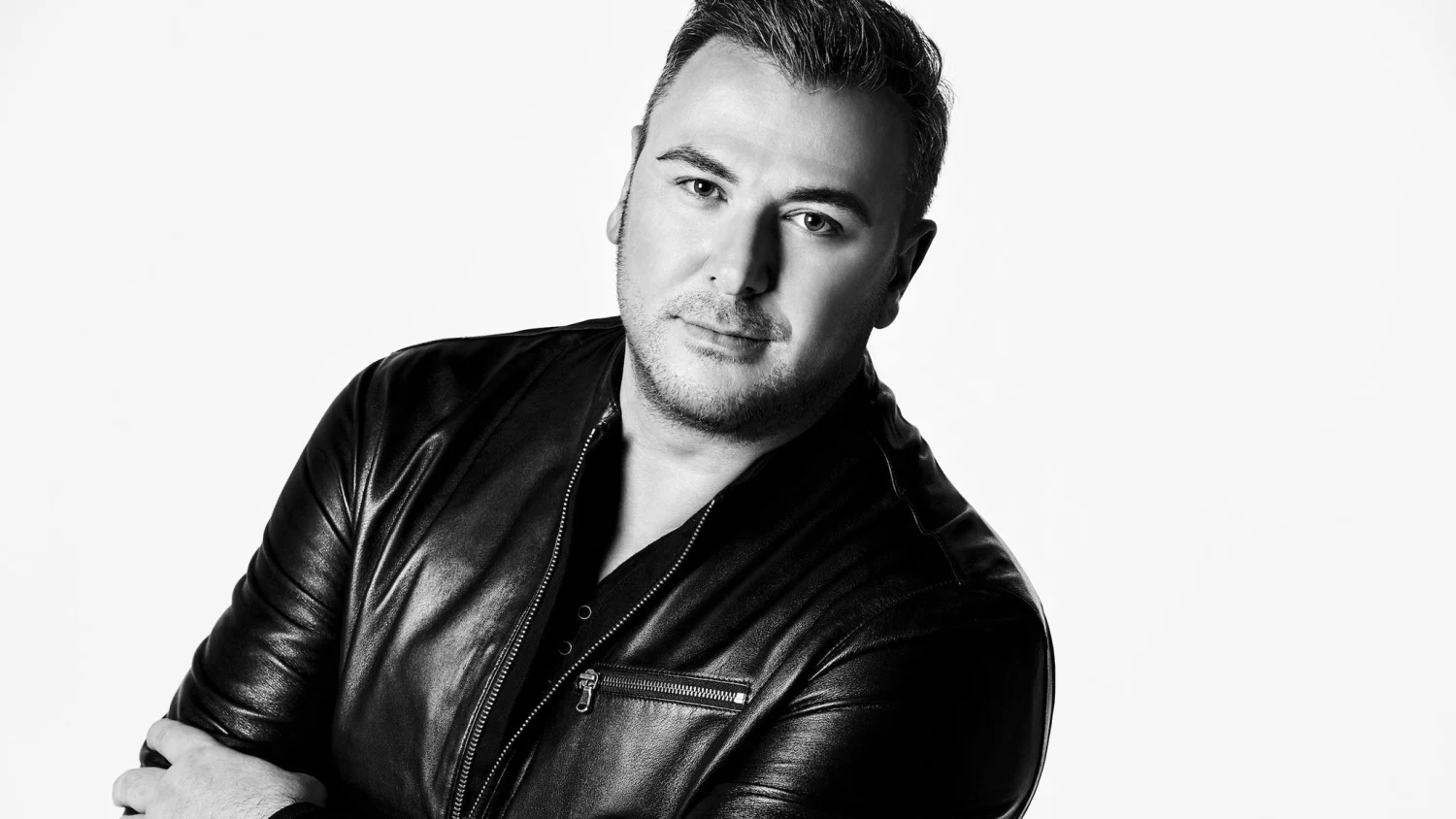 Siyah beyaz bir stüdyo fotoğrafında, ünlü şarkıcı Antonis Remos, kameraya doğrudan bakarak, hafif bir gülümsemeyle ve sakallı yüzüyle poz veriyor; parlak siyah deri ceketi önde açık, altında koyu renk bir tişört ve ceketin metal fermuar detayı dikkat çekiyor.