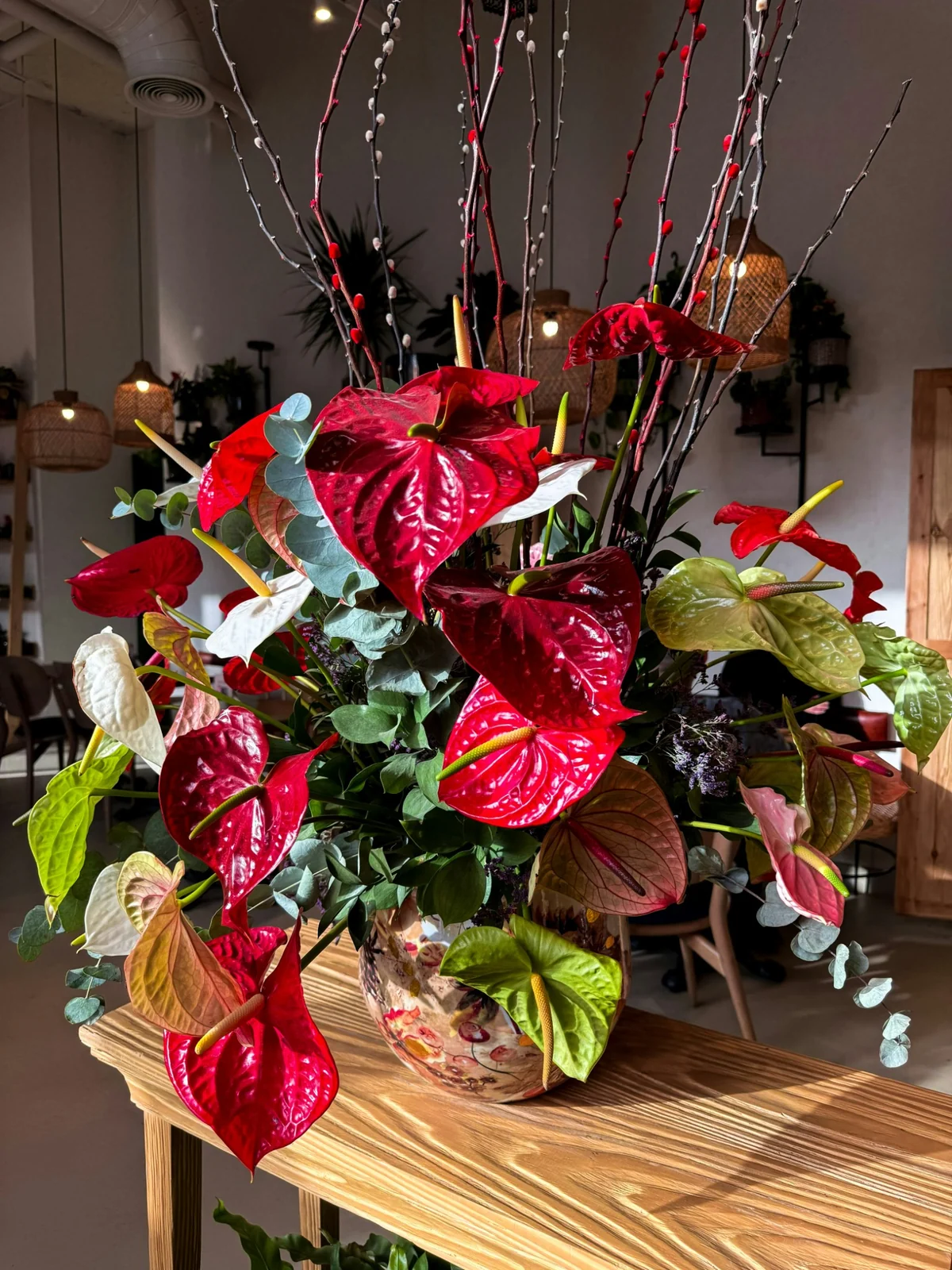 Ahşap bir masa üzerinde, parlak kırmızı, beyaz ve yeşilin farklı tonlarındaki anthurium çiçekleri, okaliptüs yaprakları, kedi söğüdü ve kırmızı tomurcuklu dallarla harmanlanarak oluşturulmuş büyük, sanatsal desenli bir vazodaki dekoratif aranjman, loş ışıklı modern bir iç mekanda büyüleyici bir atmosfer yaratıyor.