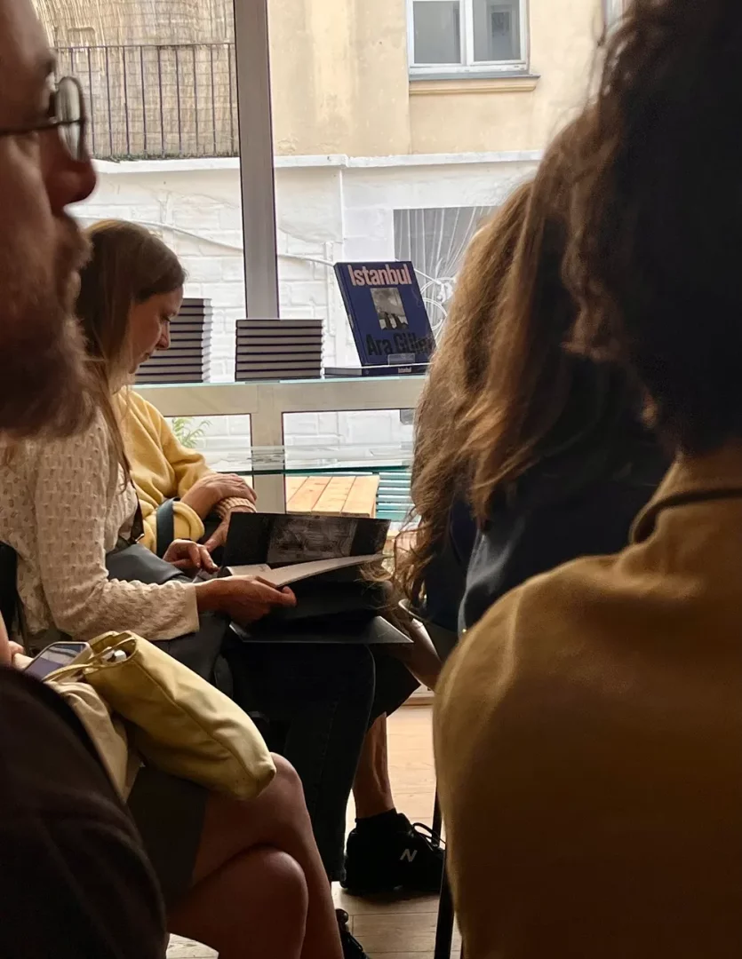 Bir etkinlik veya sunum sırasında, ön plandaki izleyicilerin başları ve omuzları görülürken, ortada cam bir masanın üzerinde efsanevi fotoğrafçı Ara Güler'in İstanbul temalı mavi kitabı, yanında yığılmış diğer yayınlarla birlikte ilgi odağı olarak duruyor.