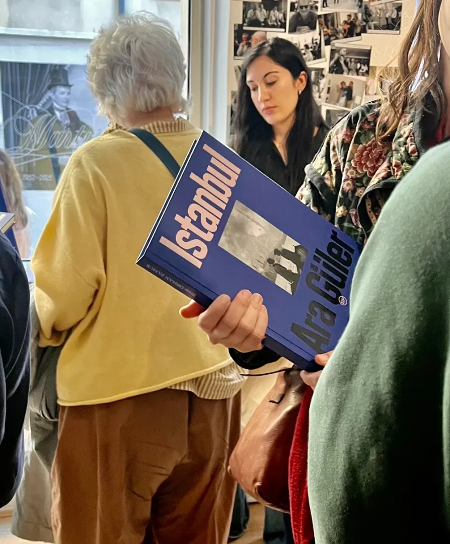 Bir kişi, arka planda fotoğraf sergisi veya kitap etkinliği kalabalığı içinde, kapağında İstanbul ve Ara Güler yazan, siyah beyaz fotoğraf baskılı mavi bir kitap tutuyor, bu görüntü Ara Güler'in Fransa'daki sanatsal etkileşimini yansıtan bir anı sunuyor.