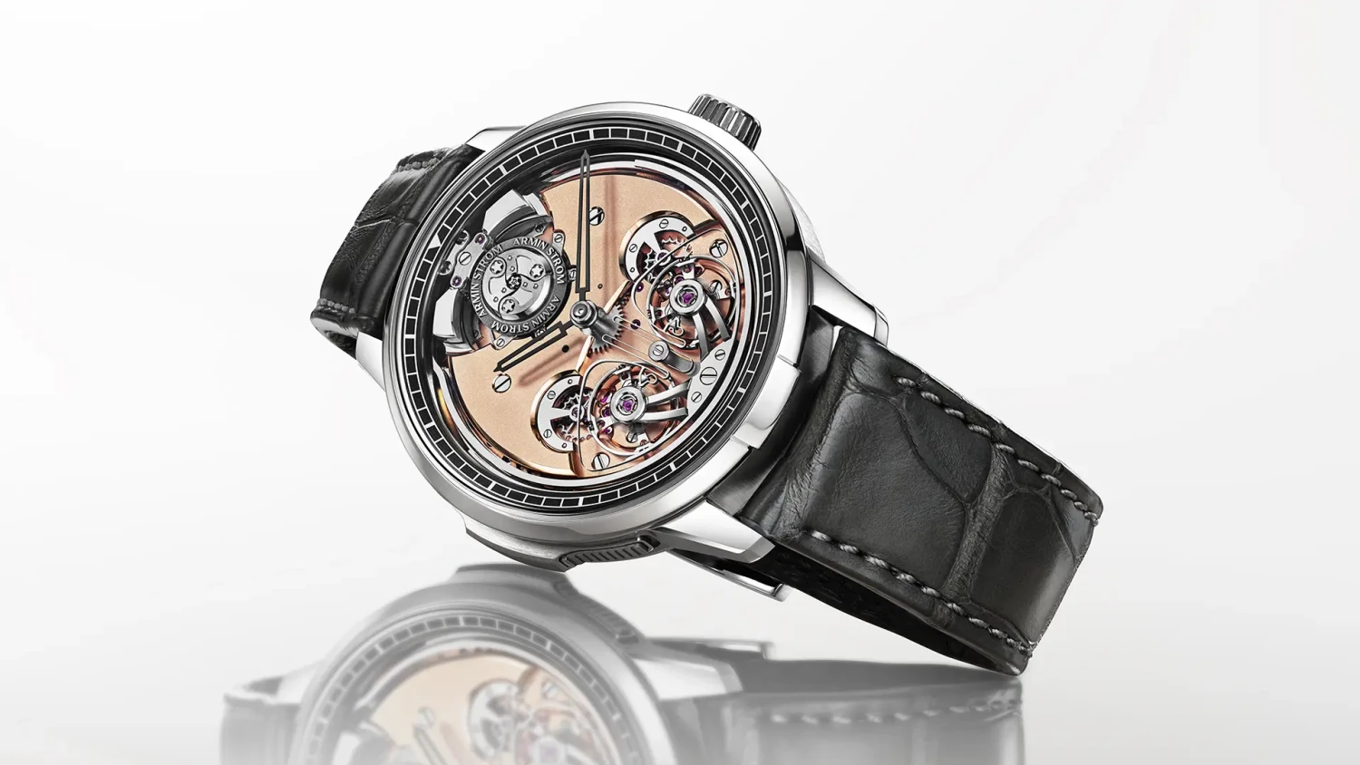 Parlak beyaz bir zeminde hafifçe eğik duran, gümüş rengi kasalı, siyah timsah derisi kayışlı ve rose gold tonlarındaki iskelet kadranında karmaşık çarklar ile minute repeater ve resonance mekanizmalarını sergileyen bir Armin Strom kol saati.