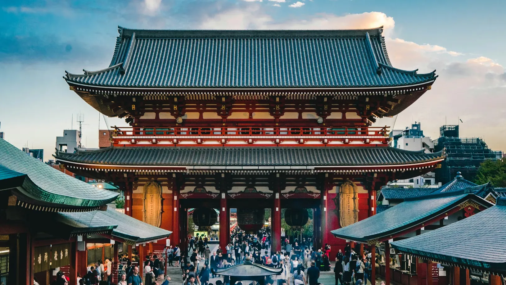 Geleneksel Japon mimarisine sahip Asakusa'daki görkemli bir tapınağın önünde, akşam saatlerinde yoğun insan kalabalığı, karmaşık kırmızı detaylar ve koyu renkli kiremit çatılar altında toplanmış, hareketli bir sahne oluşturuyor.
