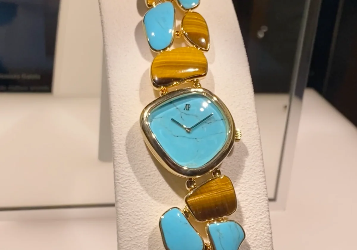 Beyaz bir stantta sergilenen, Audemars Piguet Galets koleksiyonundan turkuaz kadranlı ve turkuaz ile kaplan gözü taşlarından oluşan pürüzsüz, düzensiz şekilli bileziğe sahip altın renkli lüks bir saat, zarif tasarımıyla Watches and Wonders etkinliğindeki yenilikleri yansıtıyor.