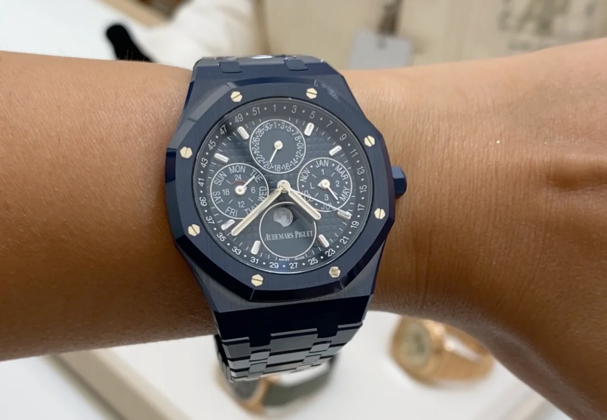 Bir bilekte duran Audemars Piguet Royal Oak Sonsuz Takvim saati, mavi seramik kasası ve kadranındaki karmaşık alt göstergeleriyle dikkat çekiyor; bu göstergeler haftanın günlerini, ayları, tarihleri ve ay evrelerini net bir şekilde sergiliyor.