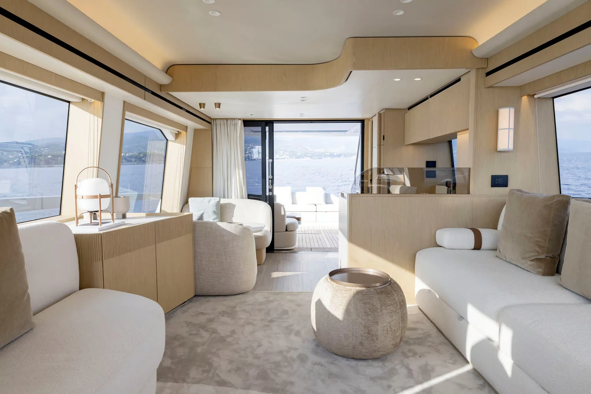 Azimut Fly 62 yatının ferah ve modern iç mekanı, büyük cam pencerelerden görünen deniz manzarasıyla birleşen açık renk ahşap panelleri, krem rengi konforlu oturma grupları ve arka güverteye açılan sürgülü kapısıyla şık bir yaşam alanı sunuyor.