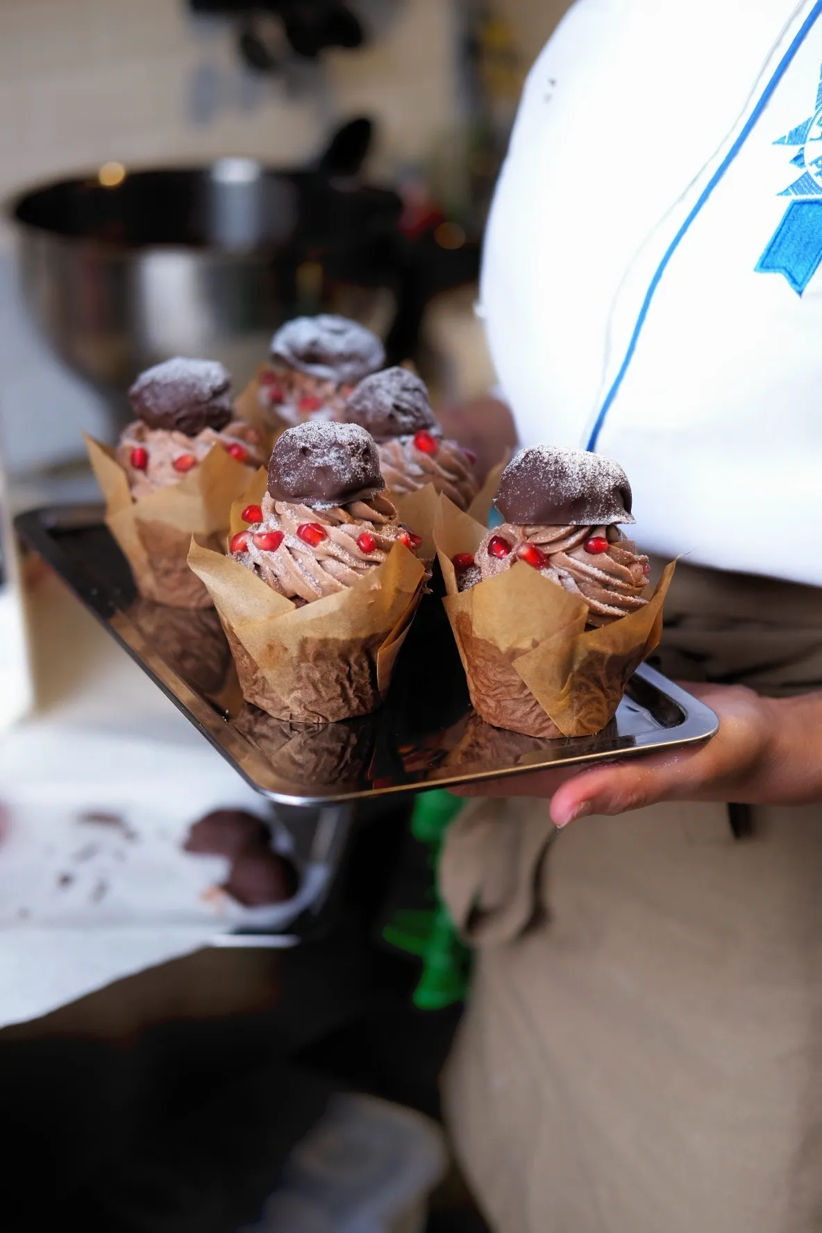 Bala Bakehouse'ta beyaz üniformalı birinin elinde, kahverengi kağıt kaplarda, çikolatalı kremayla kaplı, üzeri nar taneleri ve pudra şekerli çikolata trüfü ile süslenmiş altı enfes cupcake içeren parlak bir tepsi görülüyor.