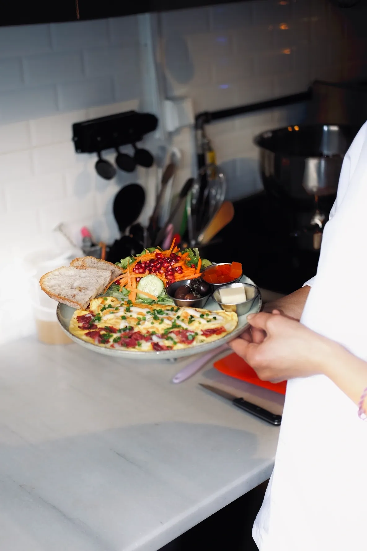 Ankara'daki Bala Bakehouse'tan beyaz önlüklü bir çalışan tarafından sunulan, sucuklu omlet, nar taneleriyle zenginleştirilmiş yeşil salata, kızarmış ekmek dilimleri, zeytin, reçel ve tereyağı içeren rengarenk ve doyurucu bir kahvaltı tabağı.