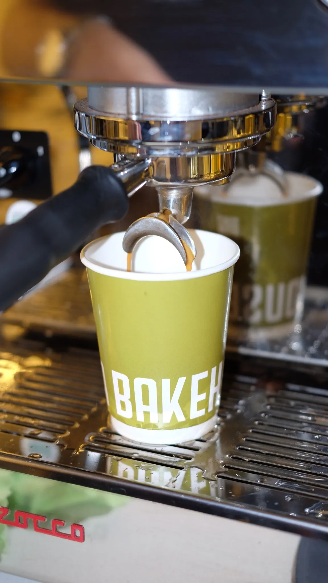 Espresso makinesinin portafiltresinden taze kahve akışı, üzerinde belirgin beyaz harflerle BAKER yazan yeşil bir kağıt bardağa dolarken, Bala Bakehouse Ankara'nın atmosferinde sıcak bir içeceğin özenle hazırlandığını gözler önüne seriyor.