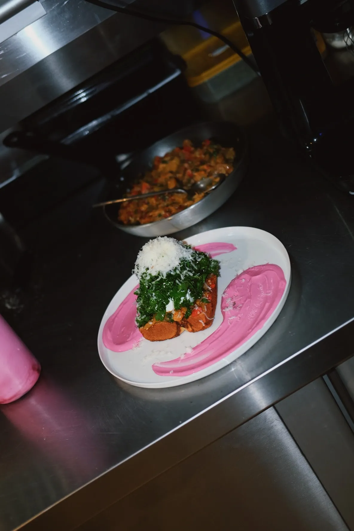 Bala Bakehouse Ankara'nın mutfak ortamını yansıtan bu karede, metal tezgah üzerinde pembe pürelerle zarifçe süslenmiş beyaz bir tabakta sunulan, tabanı kızarmış ekmek, üzeri bol yeşillik ve rendelenmiş beyaz peynirle taçlandırılmış, iştah açıcı bir brunch veya öğle yemeği tabağı dikkat çekiyor.