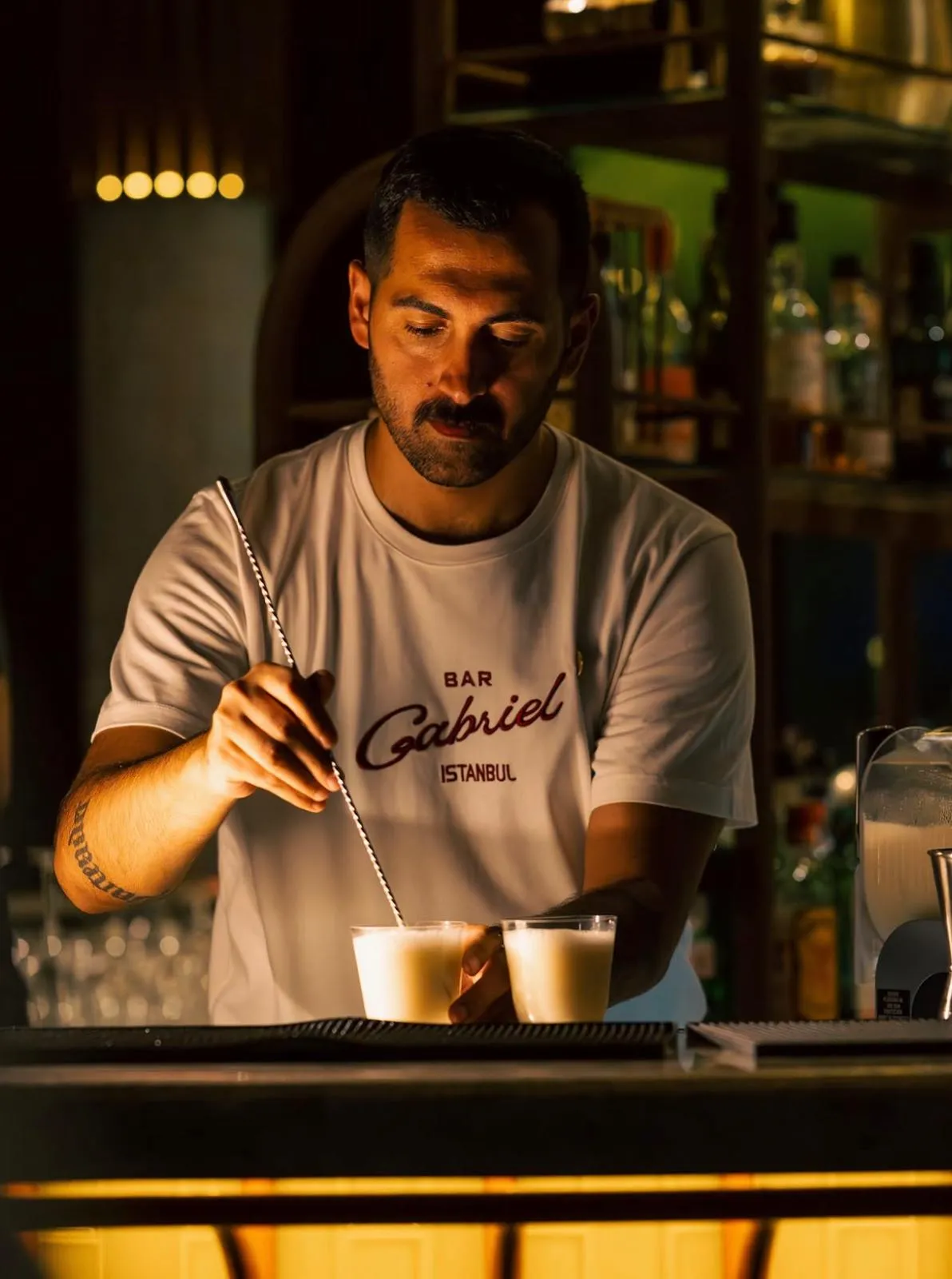 Bar Gabriel'in şık ve davetkar atmosferine sahip, loş ışıklarla aydınlatılmış modern iç mekanında, zarif bar tezgahı çevresinde oturan veya ayakta duran birkaç müşteri, mekânın özgün tasarım detayları arasında sohbet ederek içeceklerini yudumluyor.