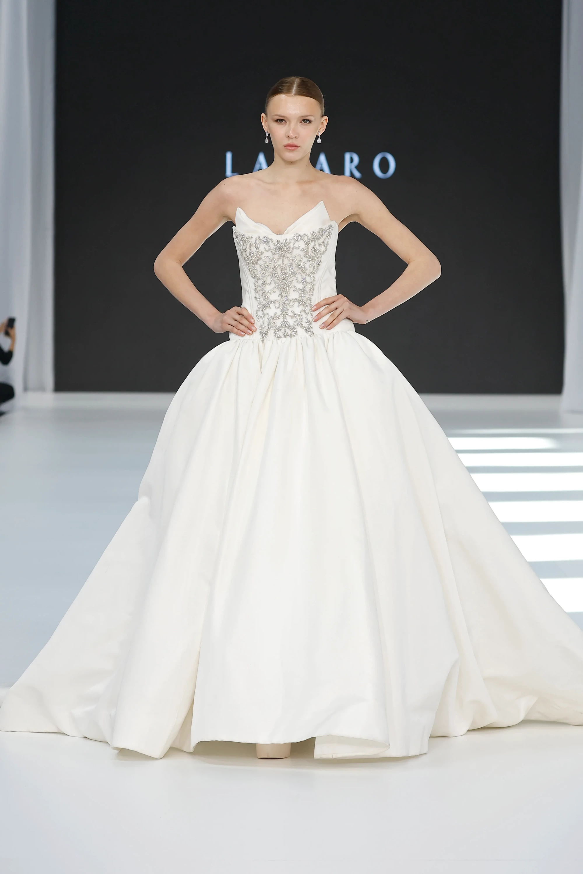 Bir podyumda elleri belinde poz veren model, straplez, gümüş renkli, zengin işlemeli korsajı ve hacimli saten etekleriyle göz kamaştıran bir Lazaro gelinlik sergiliyor, bu tasarım Barcelona Bridal Fashion Week 2026'nın öne çıkan 2027 ilkbahar gelinlik trendlerini yansıtıyor.