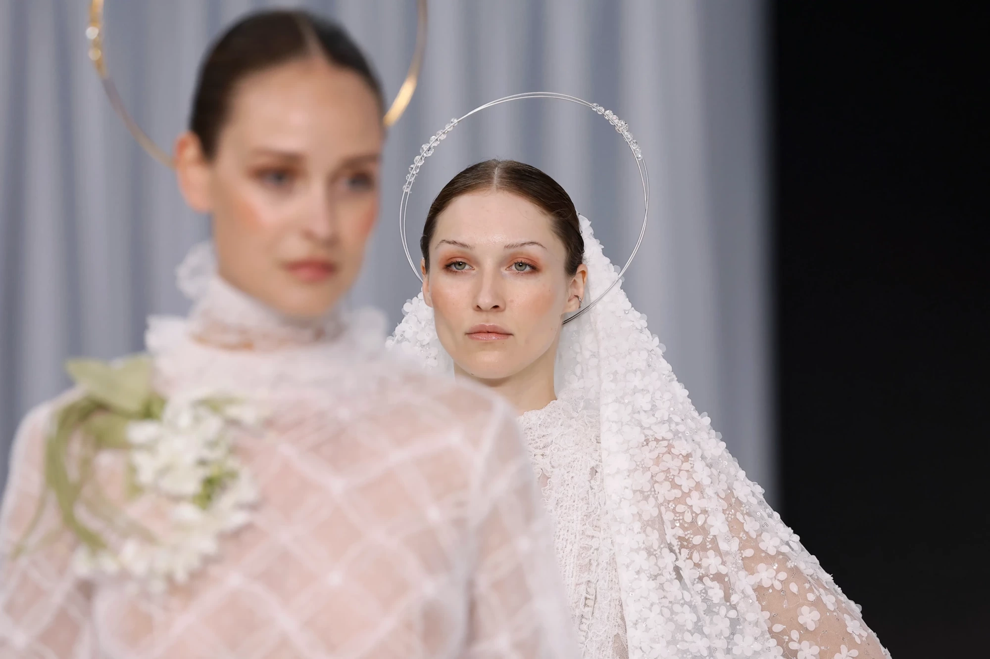 Barcelona Bridal Fashion Week 2026 podyumunda, üç boyutlu çiçek aplikeleriyle bezeli beyaz bir gelinlik giyen ve gümüş boncuklarla süslü halka başlığı takan model, ön plandaki şeffaf çiçek desenli gelinlikli modelle birlikte yeni sezon gelinlik trendlerini sergiliyor.