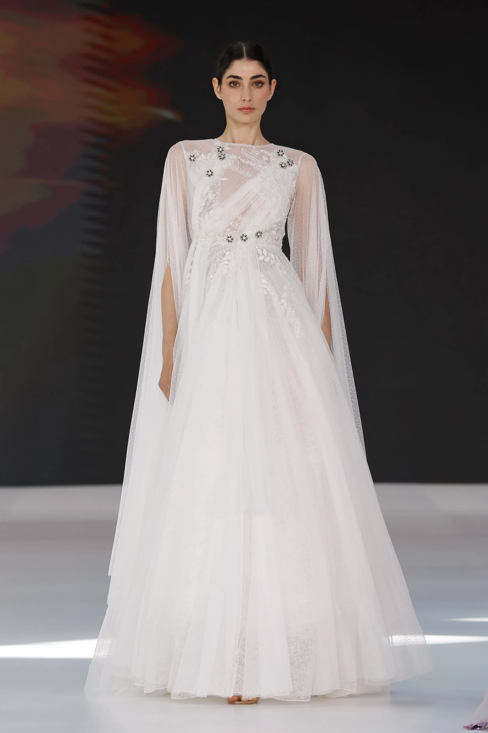 Barcelona Bridal Fashion Week 2026'da Amarca'nın 2027 ilkbahar gelinlik koleksiyonundan bir manken, transparan tül katmanları, omuzlardan zarifçe sarkan pelerin kolları ve beldeki ince çiçek aplikeleriyle romantik, akışkan ve modern bir gelinlik sunuyor.