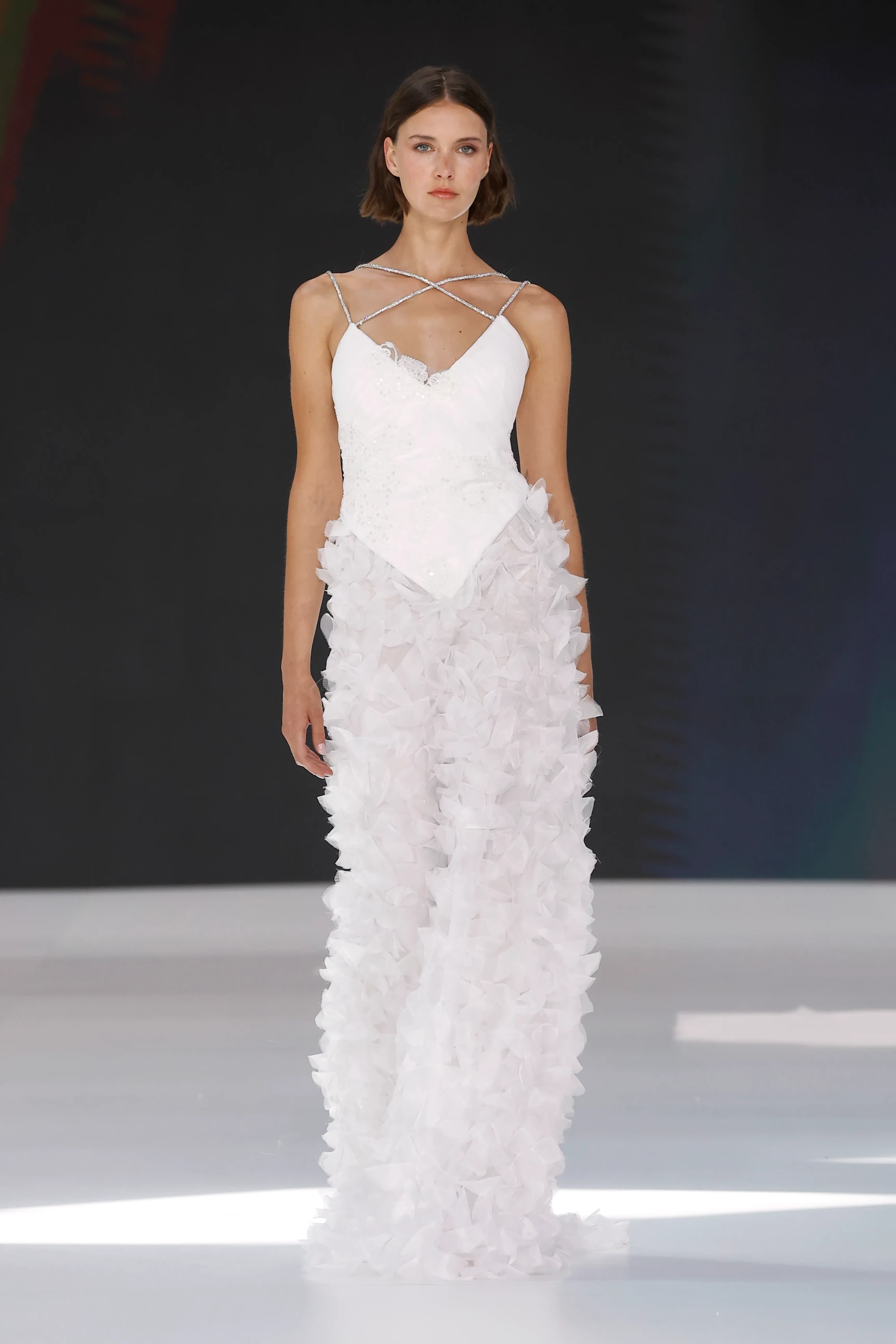 Barcelona Bridal Fashion Week'te sergilenen Amarca'nın 2027 İlkbahar Gelinlik Koleksiyonu'ndan, çapraz ışıltılı askılara sahip V yakalı korse ve üç boyutlu, hafif katmanlı tüllerle oluşturulmuş çiçek detaylı etek kısmıyla dikkat çeken beyaz bir gelinliği giyen model, podyumda duruyor.