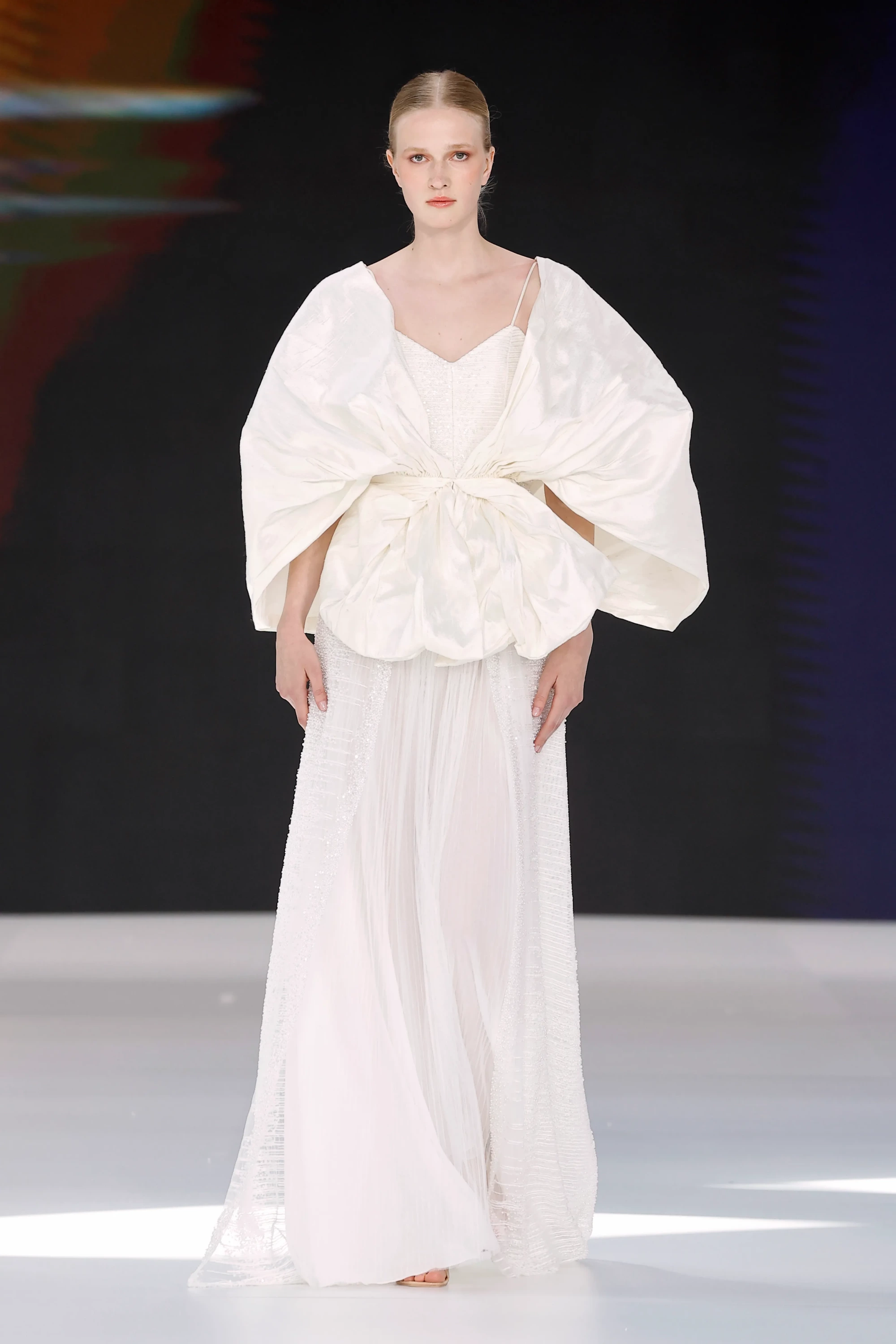 Barcelona Bridal Fashion Week 2026'da Amarca'nın 2027 İlkbahar Gelinlik Koleksiyonu'ndan podyumda sergilenen model, zarif transparan katmanları, ışıltılı işlemeleri ve üst kısmındaki heykelsi, hacimli drapeli detayları dengeleyen akışkan tül etekleriyle modern bir gelinlik sunuyor.