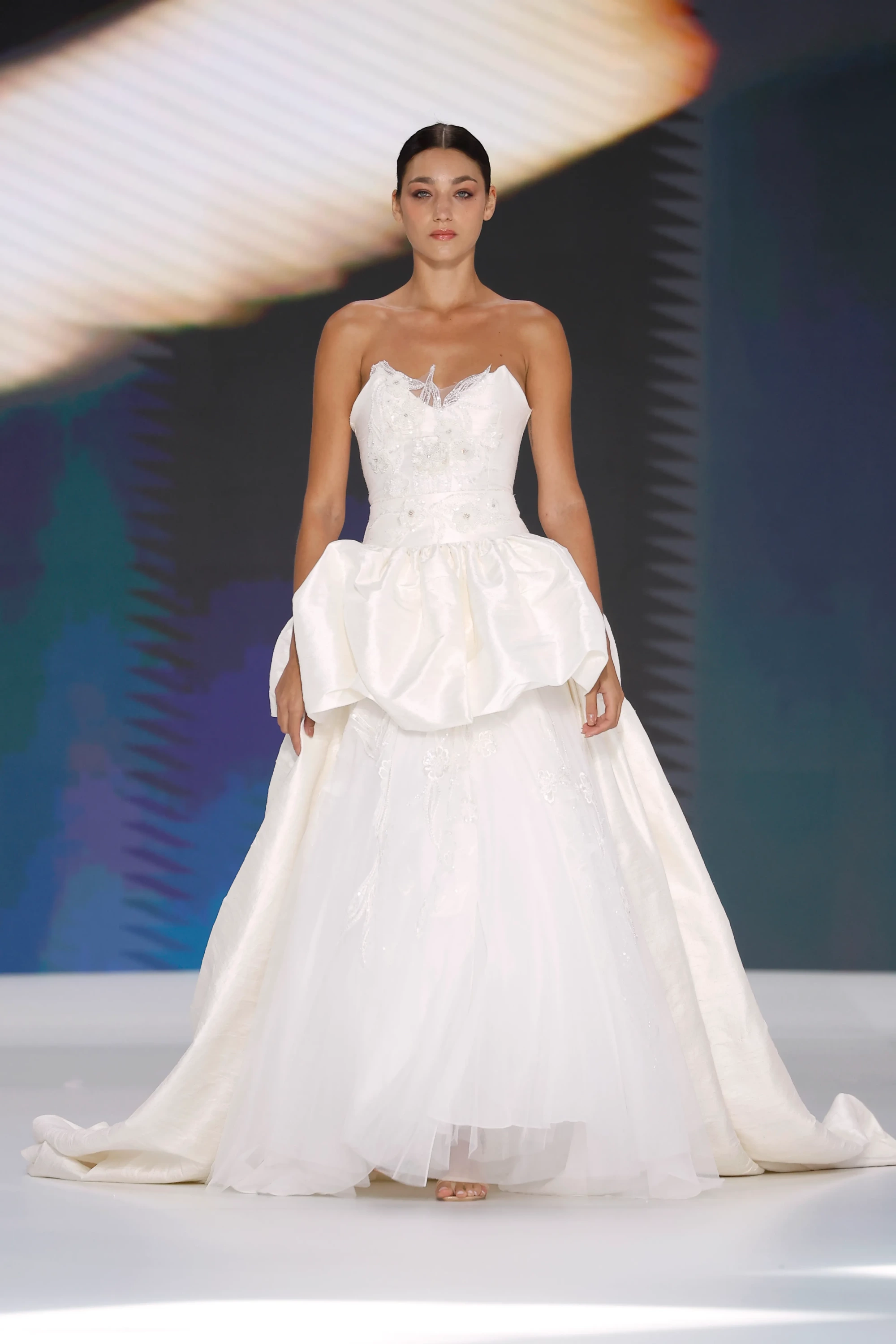 Barcelona Bridal Fashion Week 2026'da Amarca'nın 2027 İlkbahar gelinlik koleksiyonundan, straplez, çiçek işlemeli büstiyere ve belinde ipeksi, hacimli bir peplum detayına sahip, tül katmanlarla zenginleşen uzun, akışkan etekli zarif bir gelinliği tanıtan model, bu sezonun gelinlik trendlerini yansıtıyor.