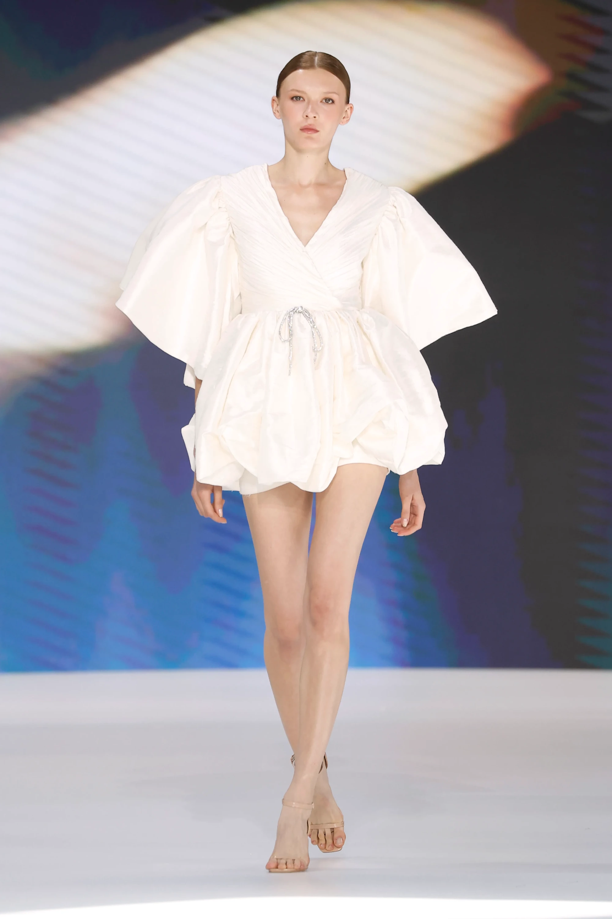 Barcelona Bridal Fashion Week 2026 kapsamında Amarca 2027 İlkbahar Gelinlik Koleksiyonu'ndan, podyumda puf kollu, pilili derin V yakalı üst kısmı ve beli ışıltılı kurdele detaylı, hacimli kısa bir gelinlik giyen model, 2027 gelinlik modasının zarif ve modern bir yorumunu sergiliyor.