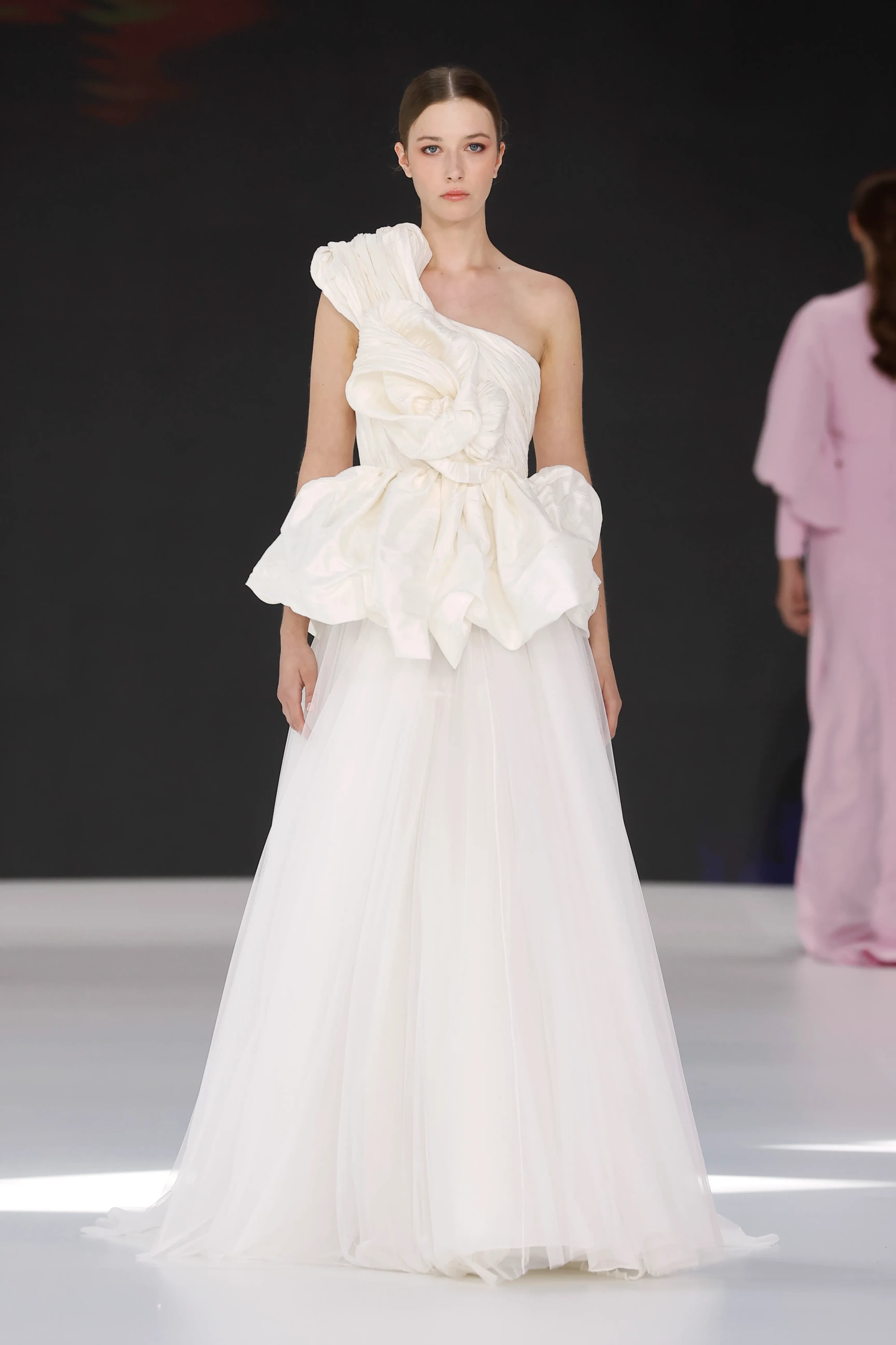 Barcelona Bridal Fashion Week 2026 podyumunda Amarca'nın 2027 İlkbahar Gelinlik Koleksiyonu'ndan, zarif ve hacimli volanlı korsajı ile dikkat çeken, omuzdan askılı ve bel kısmında büyük, çiçek benzeri fırfır detaylarıyla zenginleştirilmiş, etekleri akışkan tüllerden oluşan modern ve büyüleyici bir gelinlik modeli.