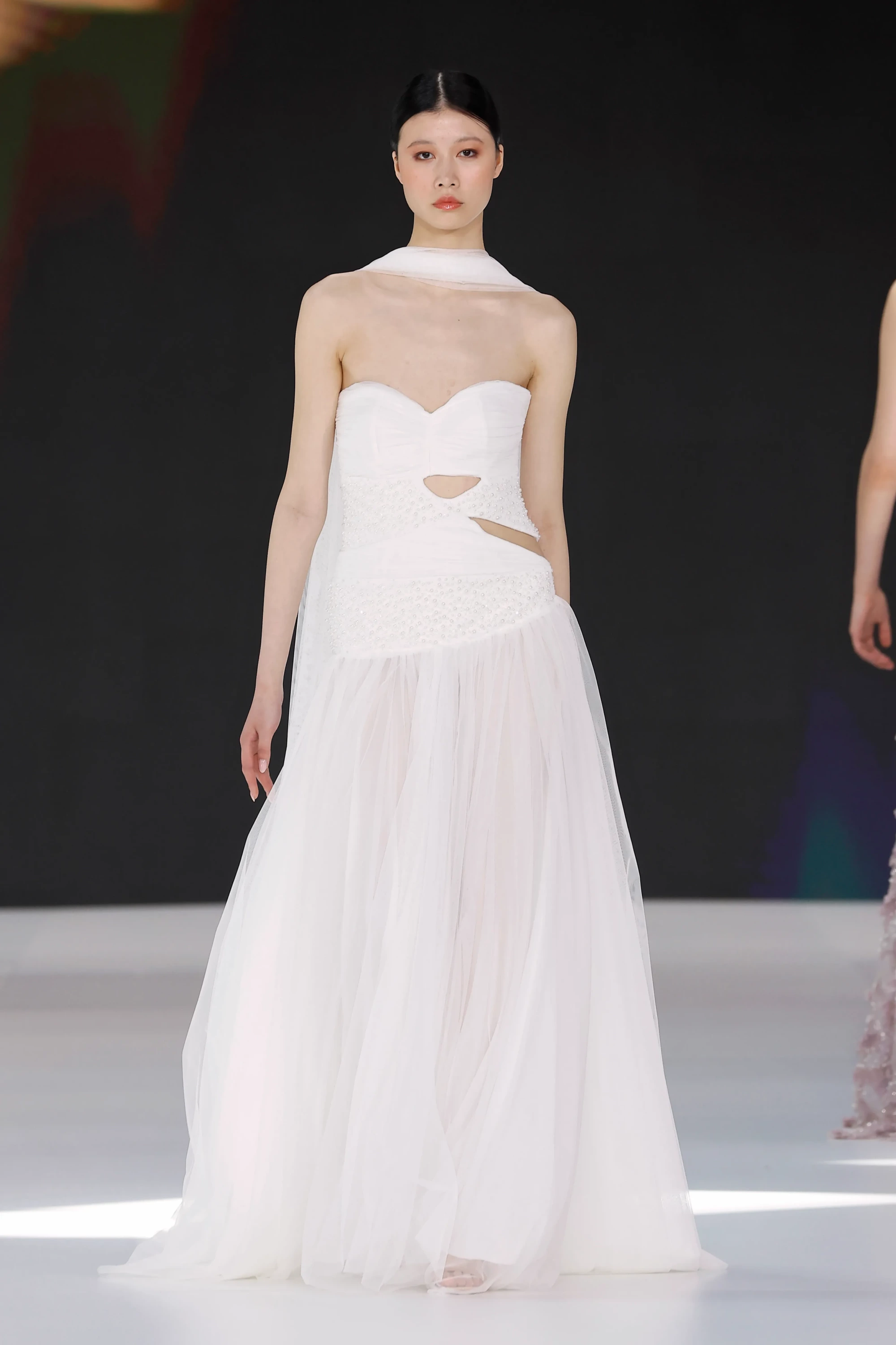 Barcelona Bridal Fashion Week 2027 İlkbahar Gelinlik Koleksiyonları'ndan Amarca tasarımı olan, yakası fular detayıyla tamamlanmış, dekolteli ve gövdesi incilerle işlenmiş, kat kat hacimli tül etekli modern bir gelinliği tanıtan Uzak Doğulu bir model podyumda duruyor.