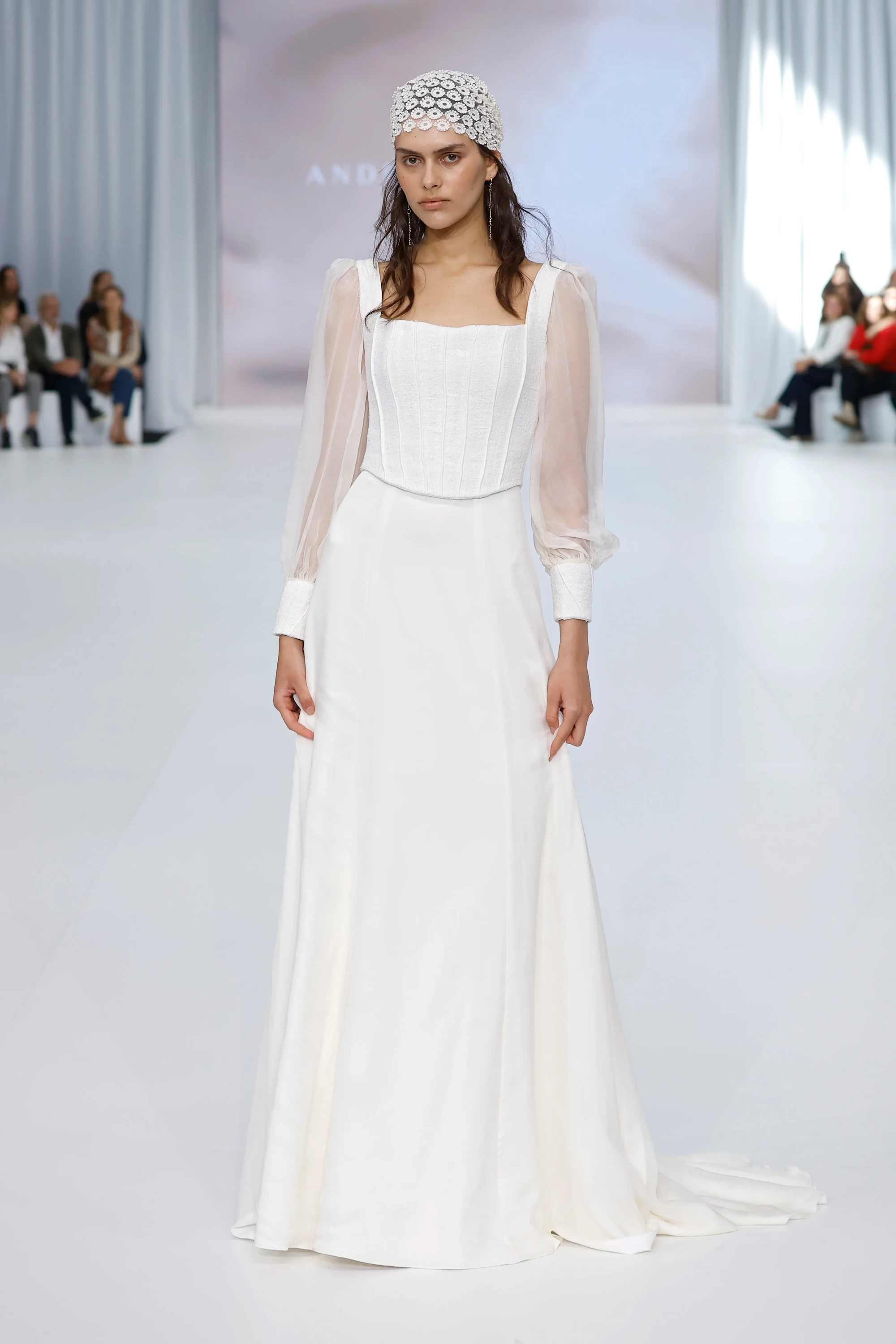 Andrea Lalanza'nın Barcelona Bridal Fashion Week 2026'daki 2027 İlkbahar gelinlik koleksiyonundan, kare yakalı korse detaylı üst kısmı ve şeffaf kollara sahip, vücutla uyumlu, sade bir gelinlik giyen model, incelikli çiçek motifli başlığıyla modern bridal zarafetini vurguluyor.