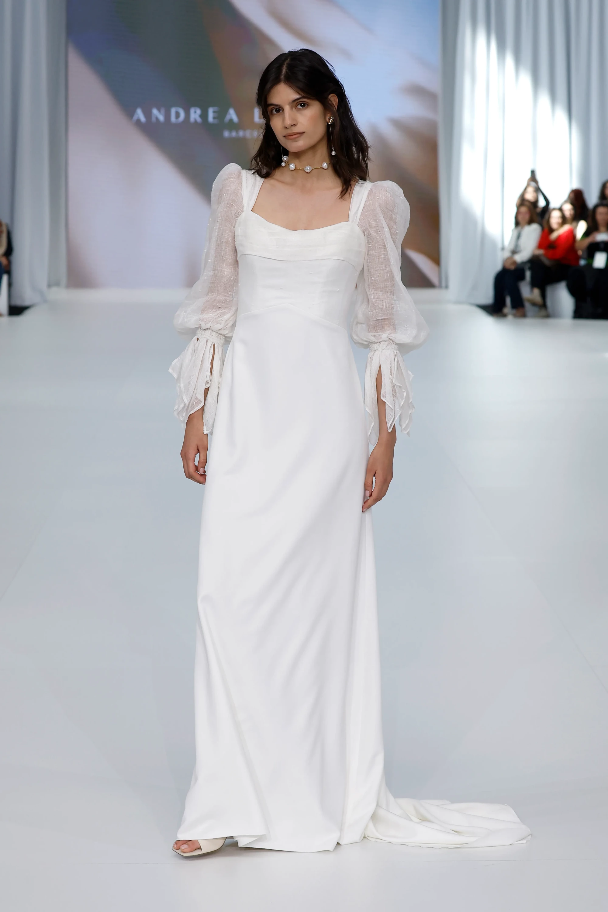Barcelona Bridal Fashion Week 2026 podyumunda, Andrea Lalanza'nın 2027 İlkbahar gelinlik koleksiyonundan, dökümlü etekli ve transparan volanlı kol detaylarına sahip beyaz, zarif bir gelinliği tanıtan model, rahat silueti ve inci aksesuarlarıyla zahmetsiz şıklığı temsil ediyor.