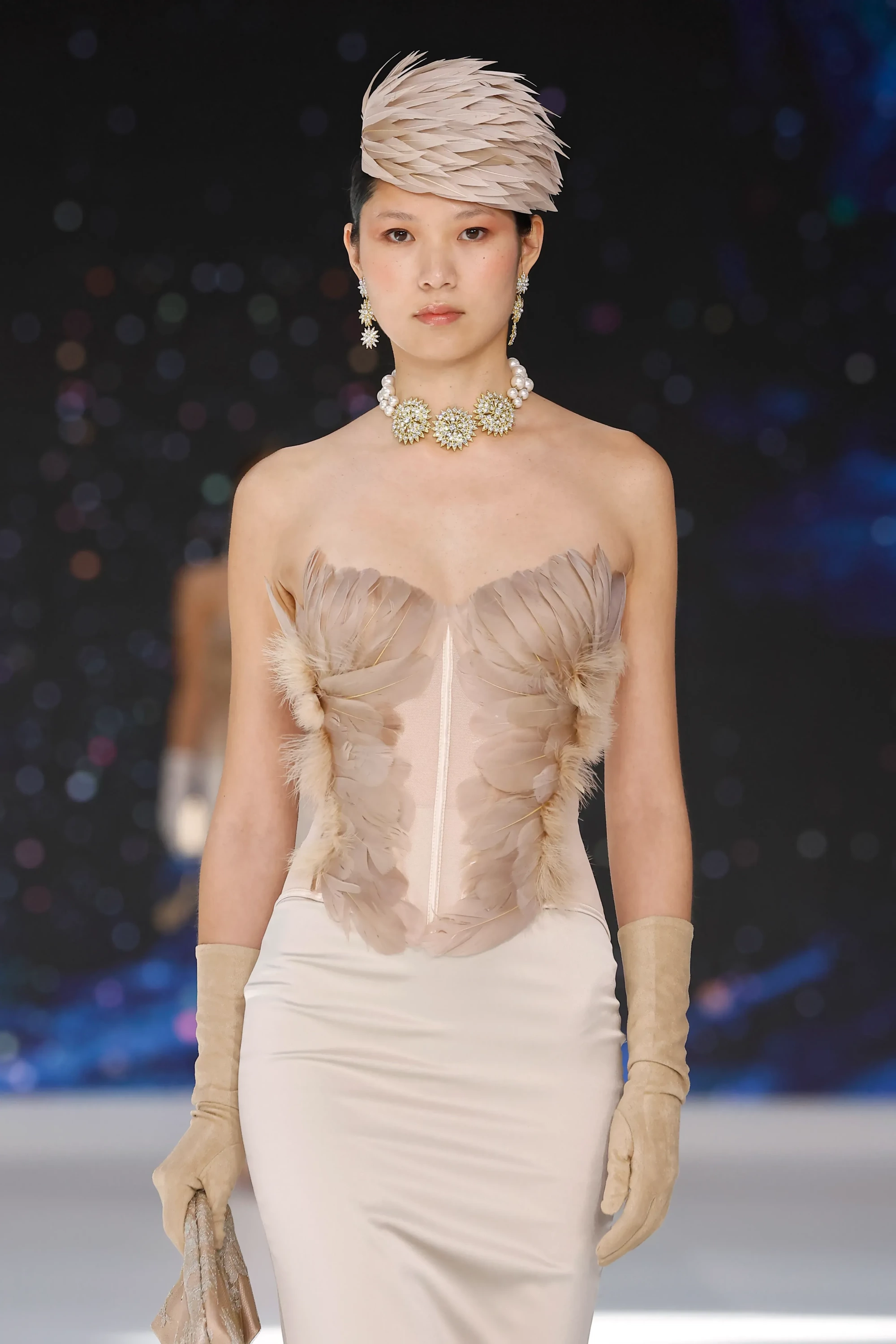 Barcelona Bridal Fashion Week podyumunda, By Loleiro 2027 İlkbahar gelinlik koleksiyonundan, tüy detaylı straplez korse üst, zarif tüy şapka, uzun eldivenler ve incili gösterişli kolyenin öne çıktığı bu görünüm, aksesuarlarla tamamlanan şık bir gelinlik trendini vurguluyor.