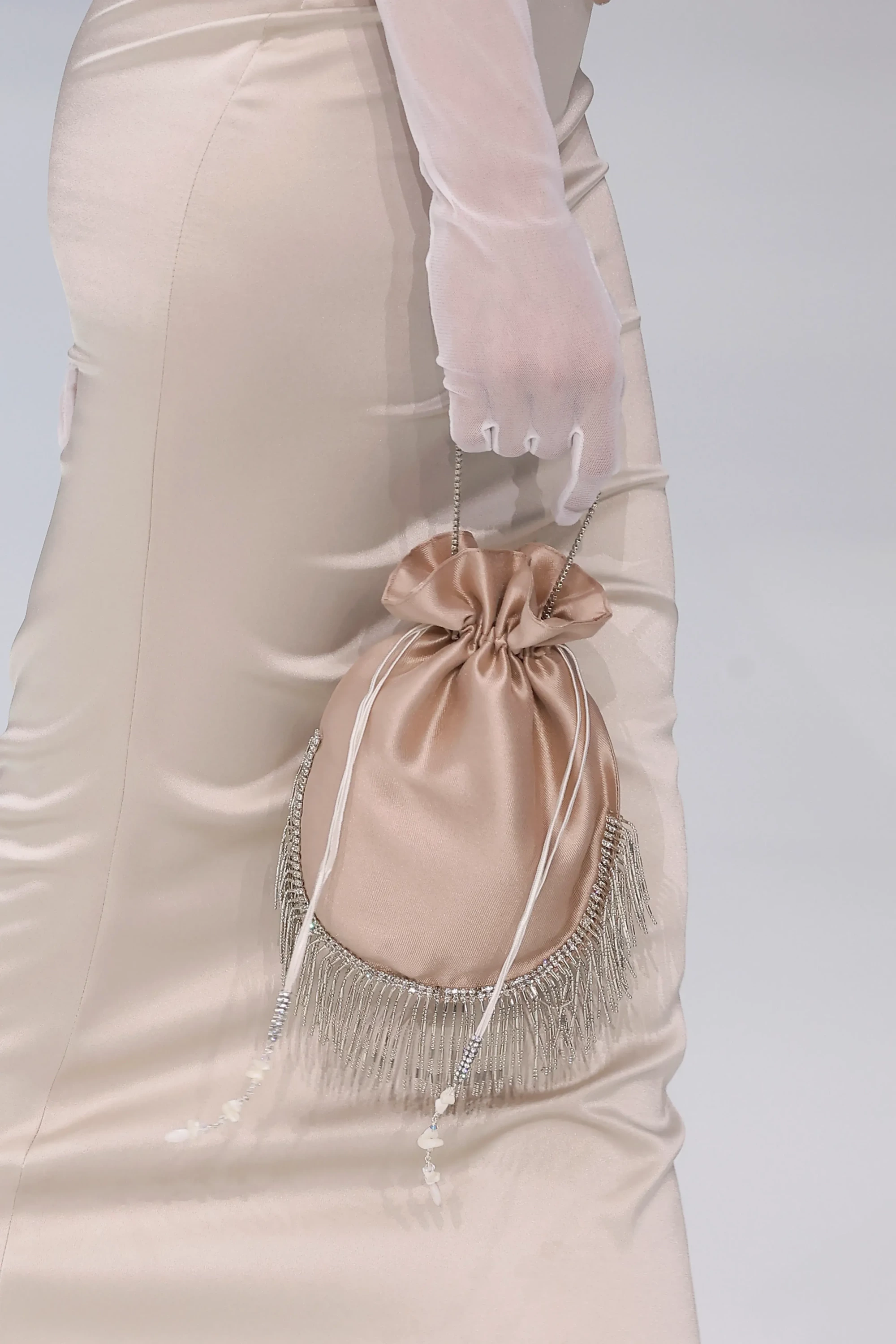 Barcelona Bridal Fashion Week'ten bir görünümde, sade, şampanya rengi saten bir gelinliğin yanında, şeffaf beyaz eldivenli bir elin tuttuğu, alt kısmı gümüş püsküller ve kristal detaylarla süslü, büzgülü, şampanya rengi saten bir çanta, By Loleiro'nun zanaatkârlık vurgusunu ve 2027 gelinlik trendlerinden aksesuarla tamamlanan gelin estetiğini sergiliyor.