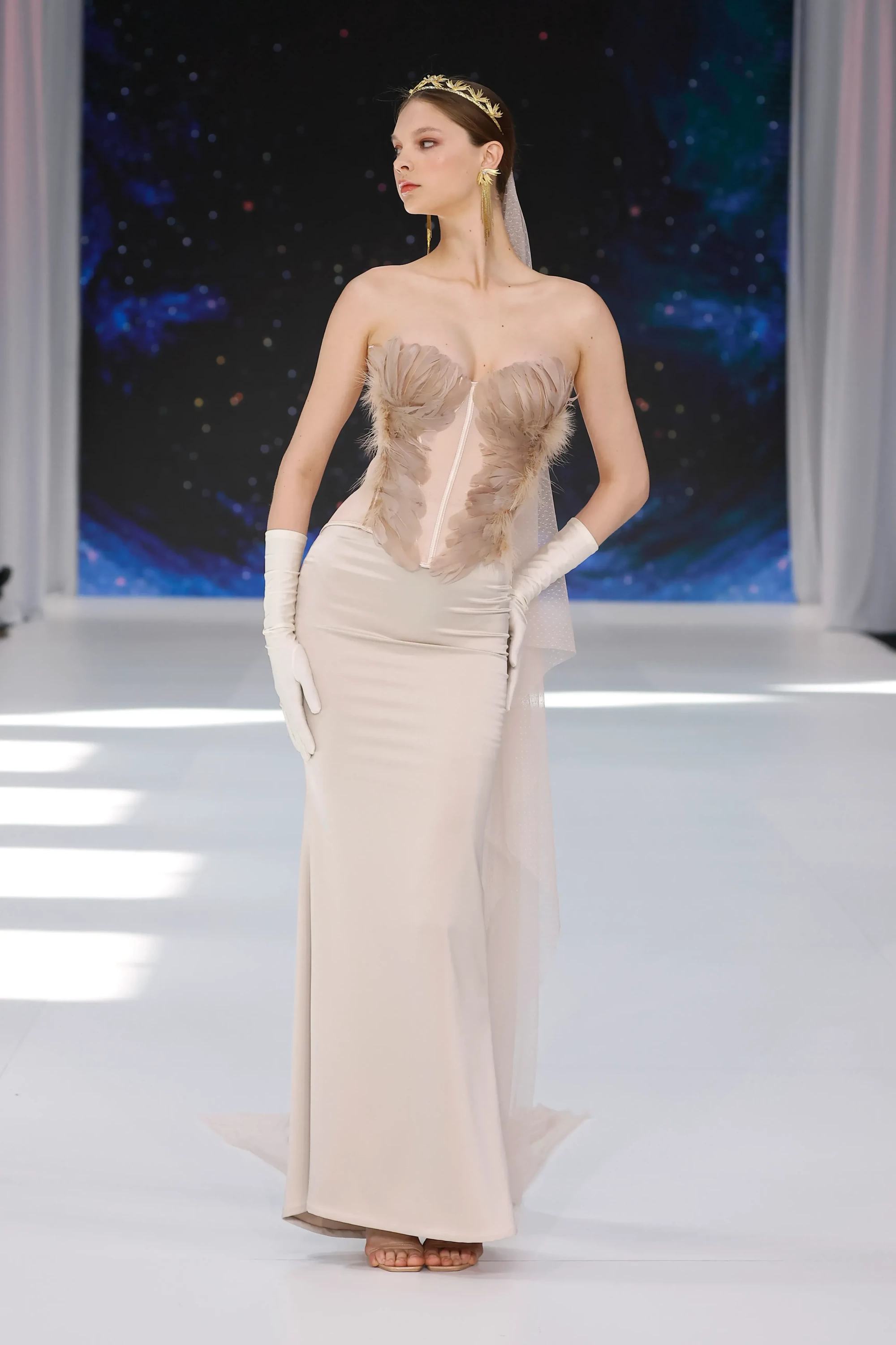 Barcelona Bridal Fashion Week 2026 etkinliğinde By Loleiro'nun Adore koleksiyonundan, tüy detaylı korseli büstiyer, uzun eldivenler, yaprak motifli altın taç ve puantiyeli tül duvak gibi aksesuarlarla stilize edilmiş, zarif, bej renkli bir gelinlik giyen model podyumda tüm bedeniyle poz veriyor.