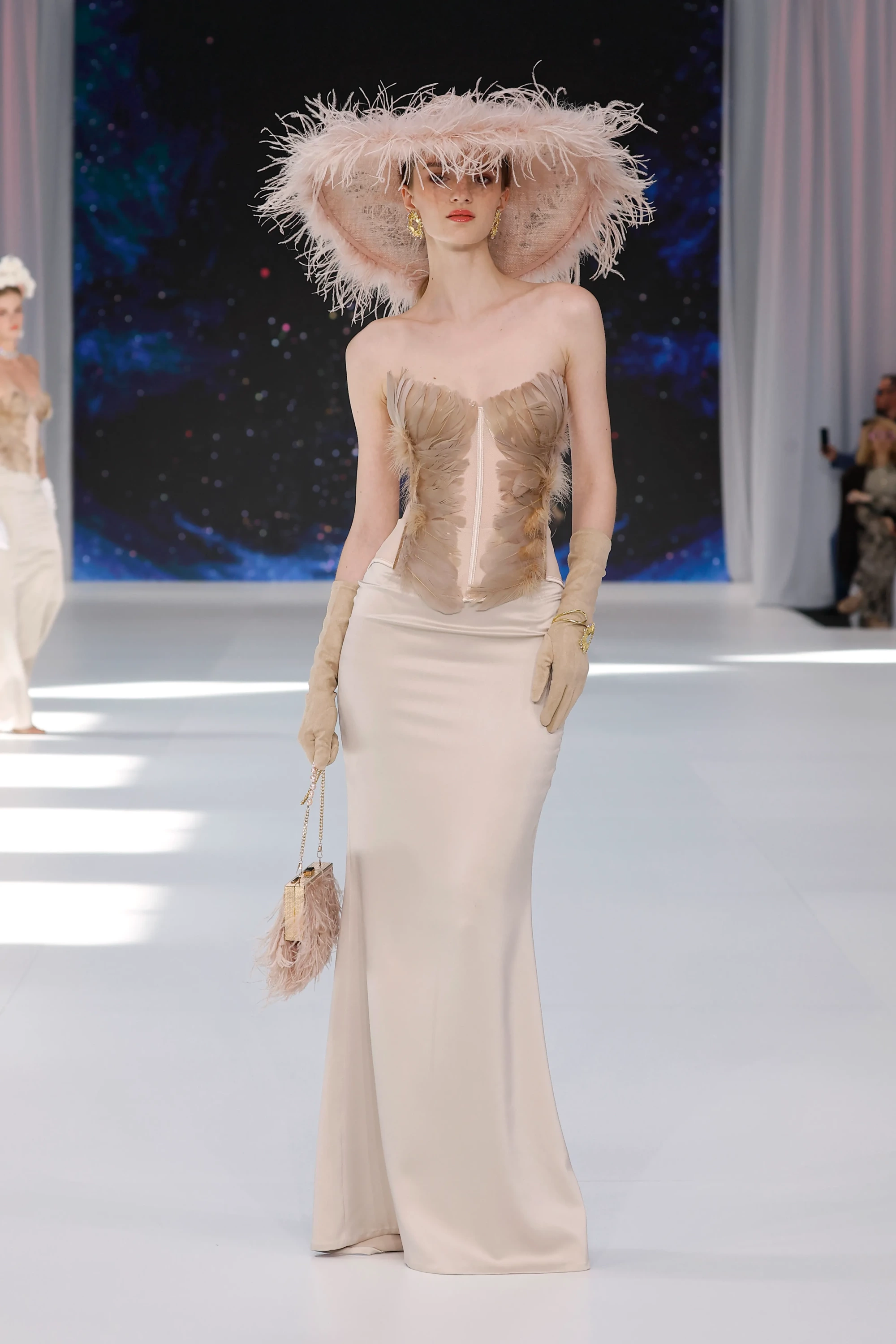 Barcelona Bridal Fashion Week 2026'da podyumda yürüyen bir model, By Loleiro'nun 2027 İlkbahar Gelinlik Koleksiyonu'ndan, tüy detaylı geniş kenarlı şapkası, bej renkli tüy süslemeli korsajı, uzun eldivenleri ve küçük tüylü çantasıyla 60'lar estetiğini yansıtan, açık bej rengi zarif bir balık etek gelinlik trendini sunuyor.