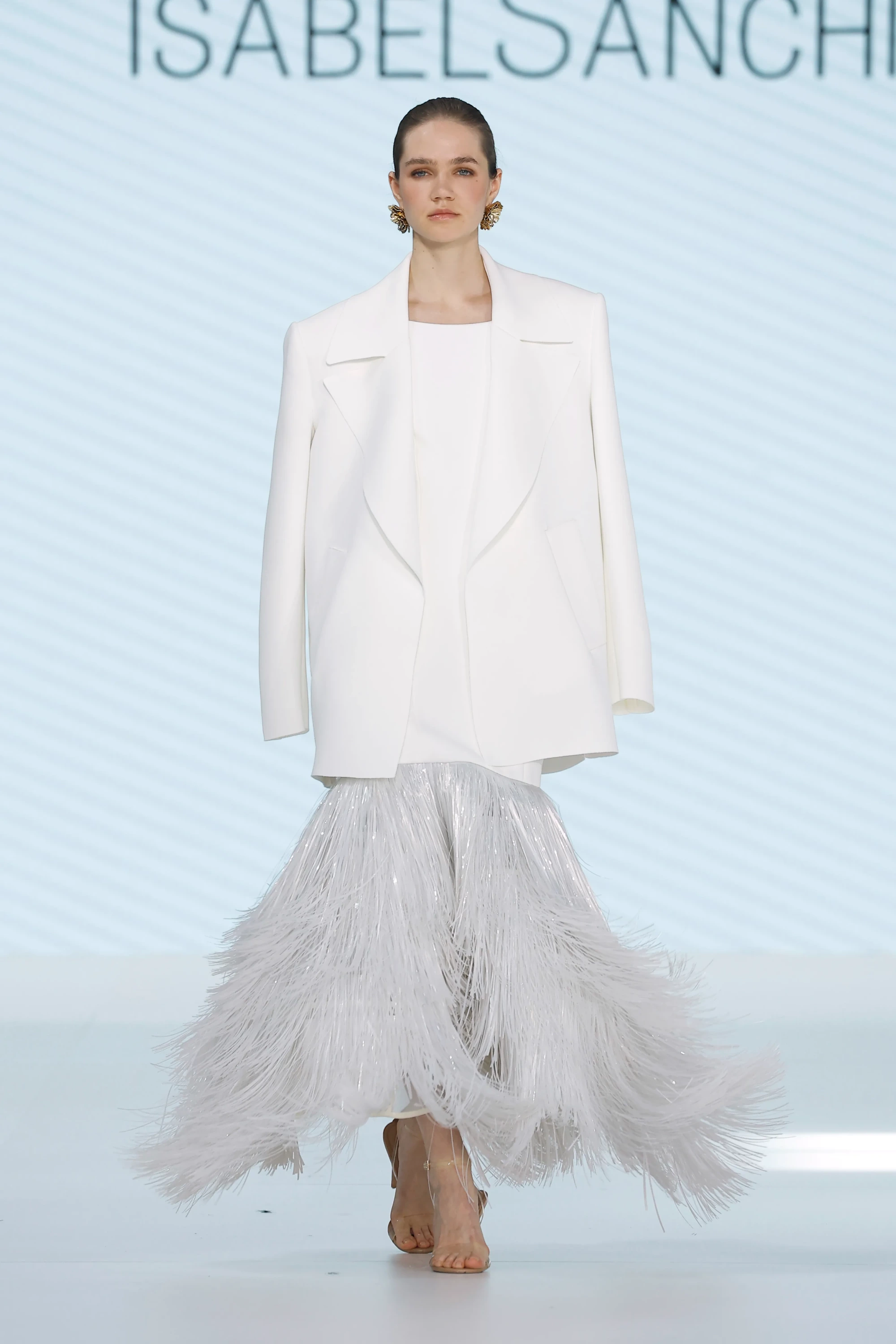 Barcelona Bridal Fashion Week podyumunda, Isabel Sanchis'in 2027 İlkbahar gelinlik koleksiyonundan beyaz, yapısal kesimli, oversized blazer ceket ile altına giyilmiş parıldayan gümüşi püsküllerden oluşan hacimli, katmanlı etekli bir gelinlik giyen model, markanın form ve mimari yaklaşımını sergiliyor.