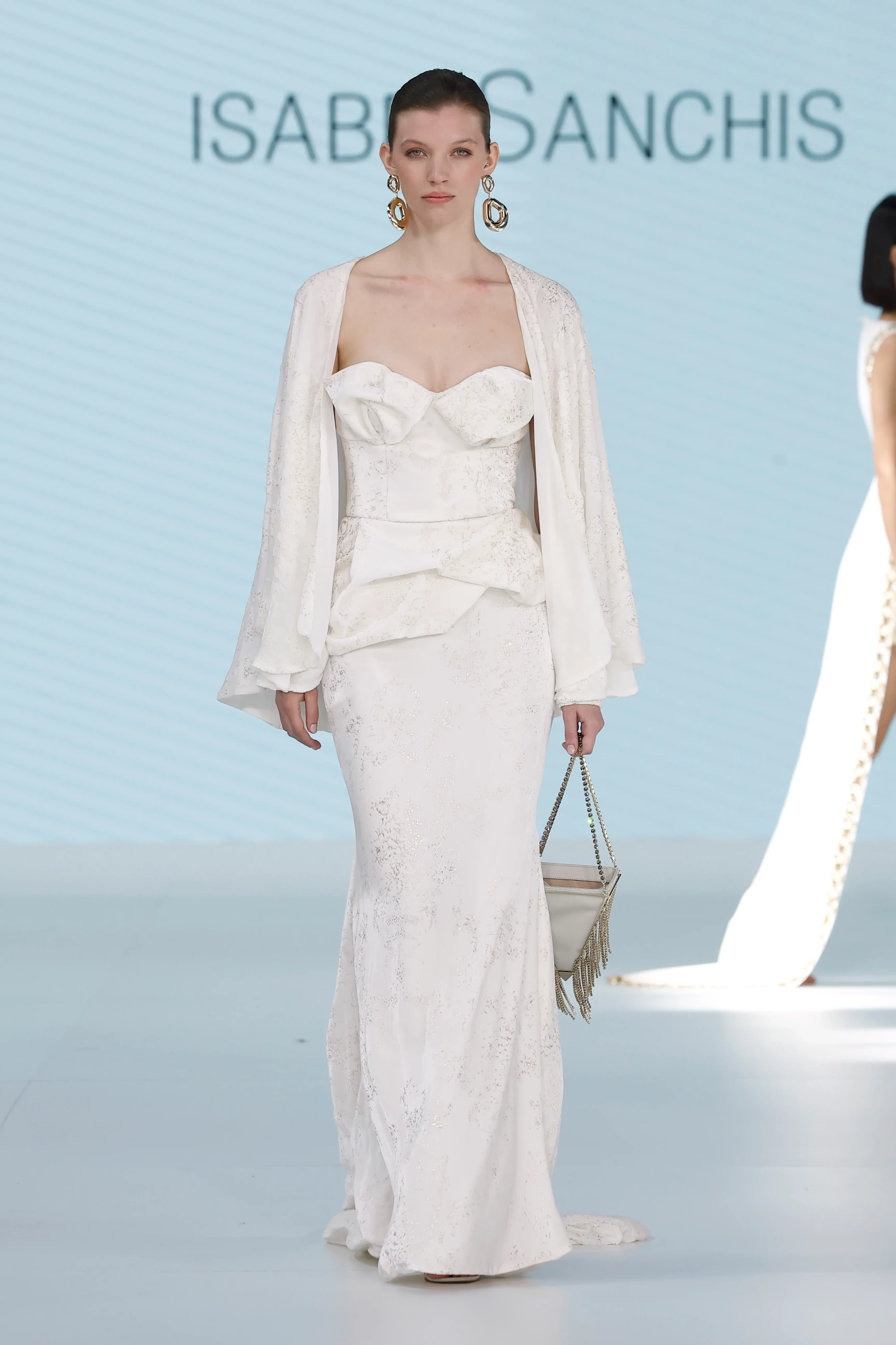 Barcelona Bridal Fashion Week 2026'dan Isabel Sanchis'in 2027 İlkbahar gelinlik koleksiyonundan bir parça sergileyen model, büstiyer kesim, belinde yapısal bir fırfır detayı olan beyaz, yerlere kadar uzanan bir elbise giyiyor. Elbiseyle uyumlu, geniş kollu, açık bir ceket, büyük altın küpeler ve püsküllü metalik bir çanta ile tamamlanan bu modern tasarım, koleksiyonun yapı ve form odaklı yaklaşımını yansıtıyor.