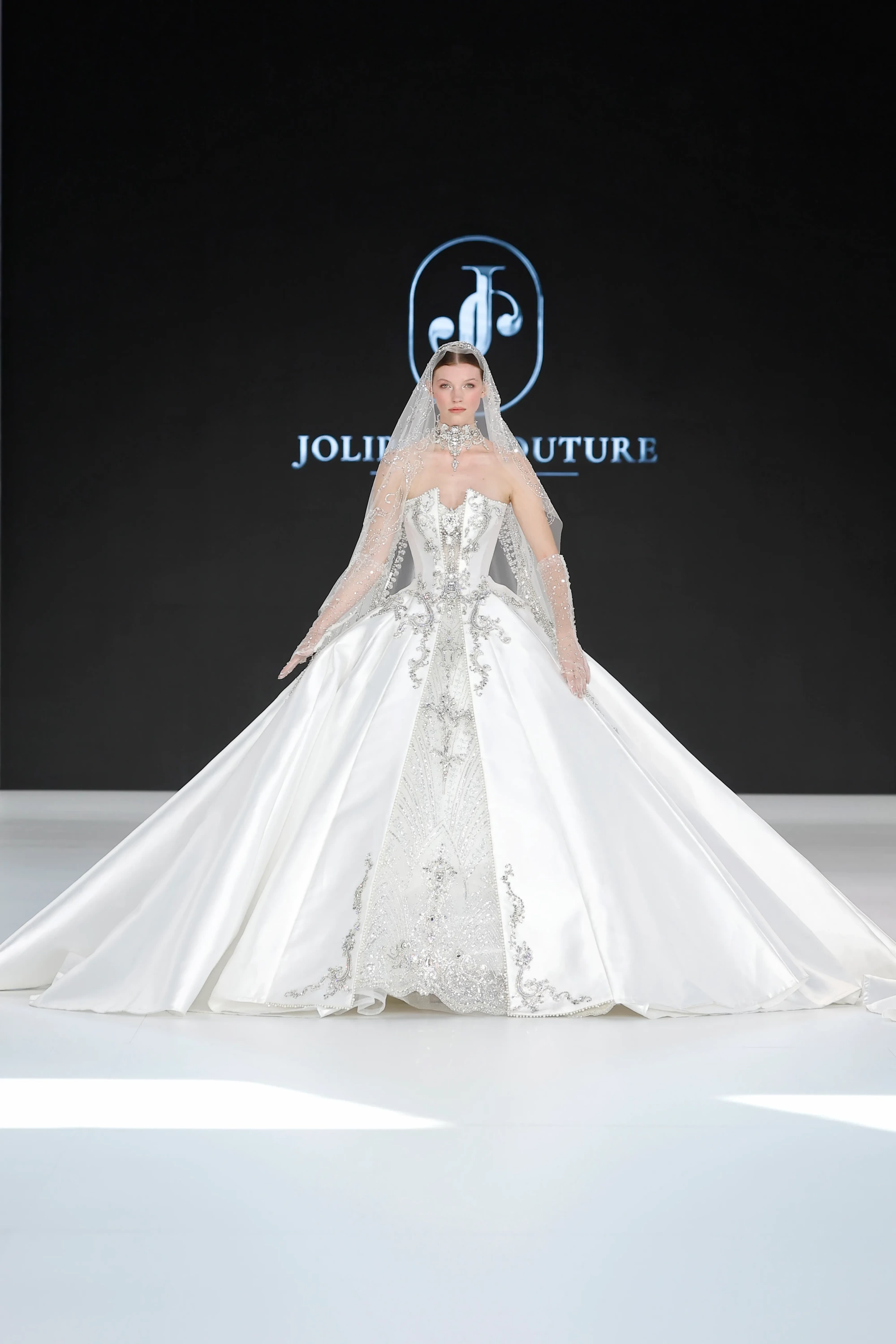 JOLI COUTURE logosunun önünde, kristal işlemelerle bezeli straplez korsajı, ihtişamlı kabarık etekleri ve uzun ışıltılı duvağıyla dikkat çeken, 2027 Gelinlik Trendleri arasında yer alan iddialı ve heykelsi bir JoliPoli gelinlik modeli podyumda duruyor.