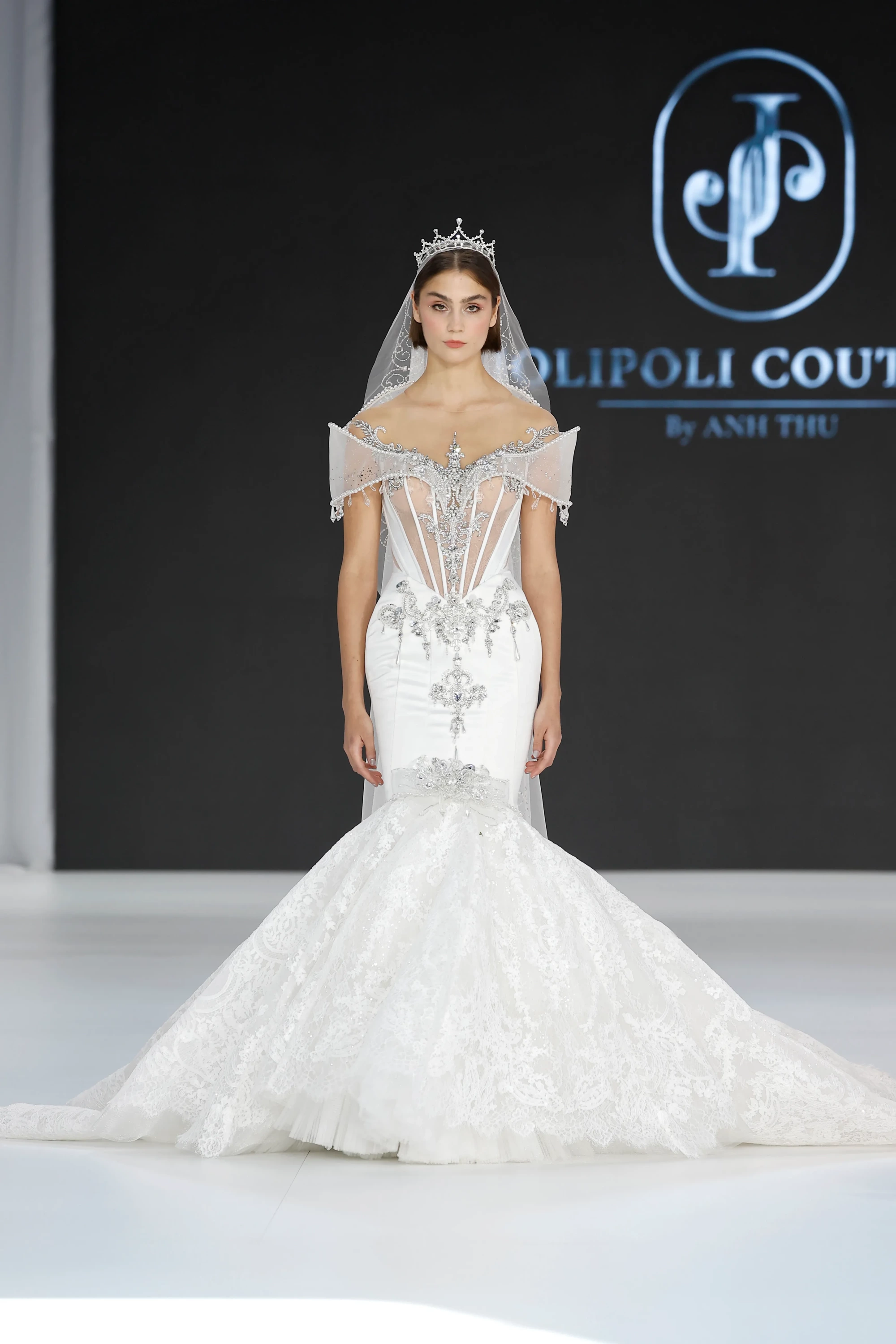 Beyaz, vücudu saran, işlemeli bir denizkızı kesim gelinlikle podyumda duran model, JoliPoli Couture imzalı, transparan korsajı ve heykelsi omuz detaylarıyla ışık saçan, dantel ve tül katmanlı kabarık eteğiyle 2027 gelinlik trendlerini yansıtan iddialı bir tasarım sergiliyor.