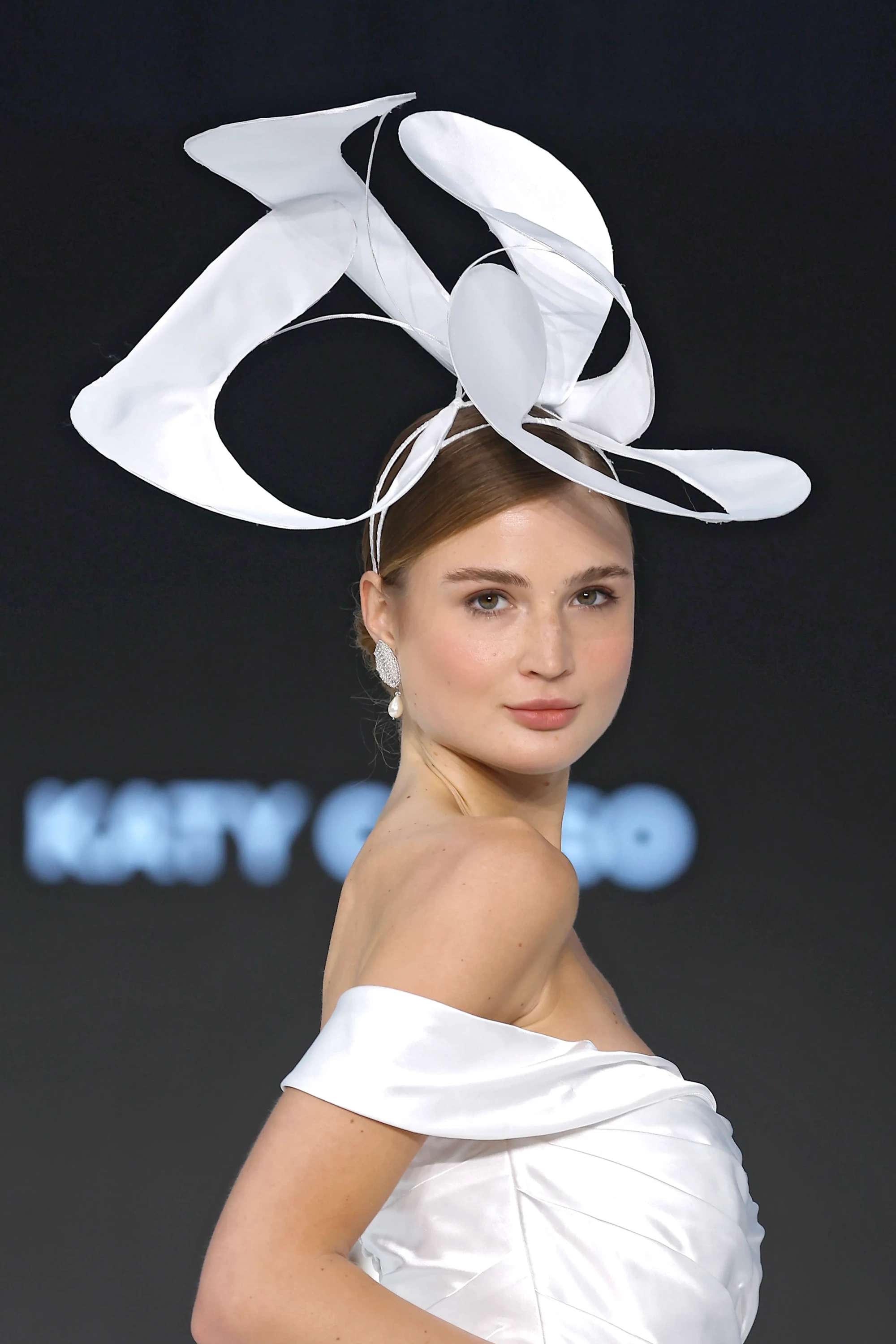 Barcelona Bridal Fashion Week kapsamında Katy Corso'nun 2027 İlkbahar gelinlik koleksiyonundan, omuzları düşük beyaz bir gelinlik giyen ve başındaki fütüristik, heykelsi beyaz aksesuarıyla zarifçe poz veren bir model.
