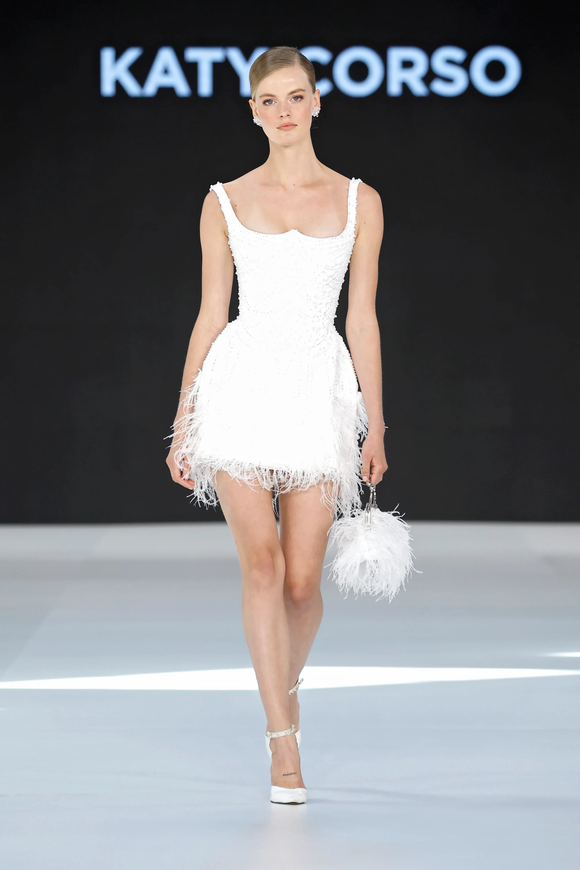 Barcelona Bridal Fashion Week 2026'da podyuma çıkan Katy Corso'nun 2027 İlkbahar gelinlik koleksiyonundan, inci işlemeli korsajı ve tüy detaylı mini eteğiyle hafiflik ve modernliği birleştiren beyaz bir gelinlik modeli, aynı tüy detaylı el çantası ve zarif topuklu ayakkabılarıyla sergileniyor.