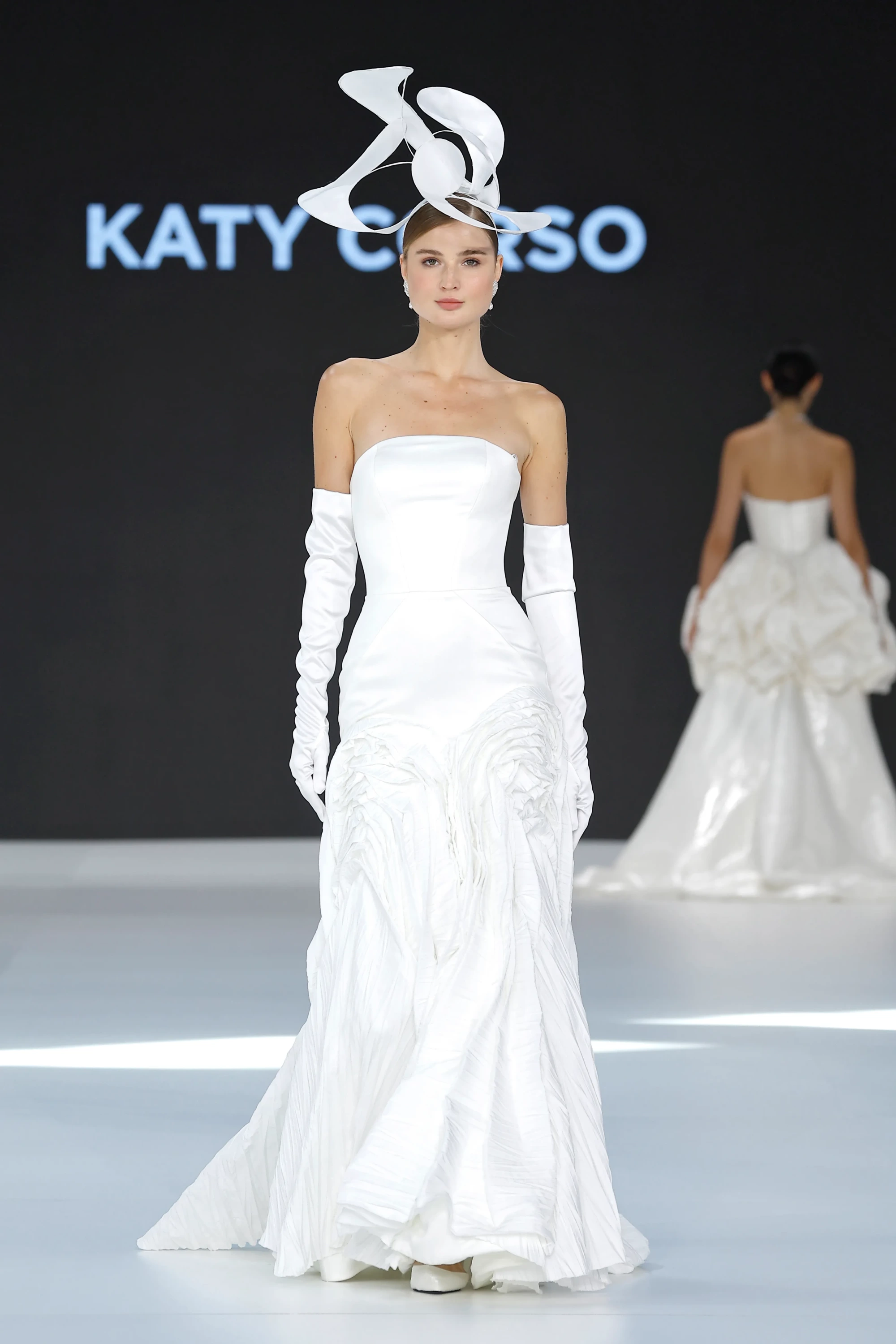 Katy Corso İlkbahar 2027 gelinlik koleksiyonundan bir model, straplez kesimli, uzun beyaz eldivenlerle tamamlanmış, etek kısmında heykelsi pilili ve büzgülü detaylara sahip modern bir gelinliği, zarif ve kıvrımlı beyaz bir baş aksesuarıyla podyumda sergiliyor.